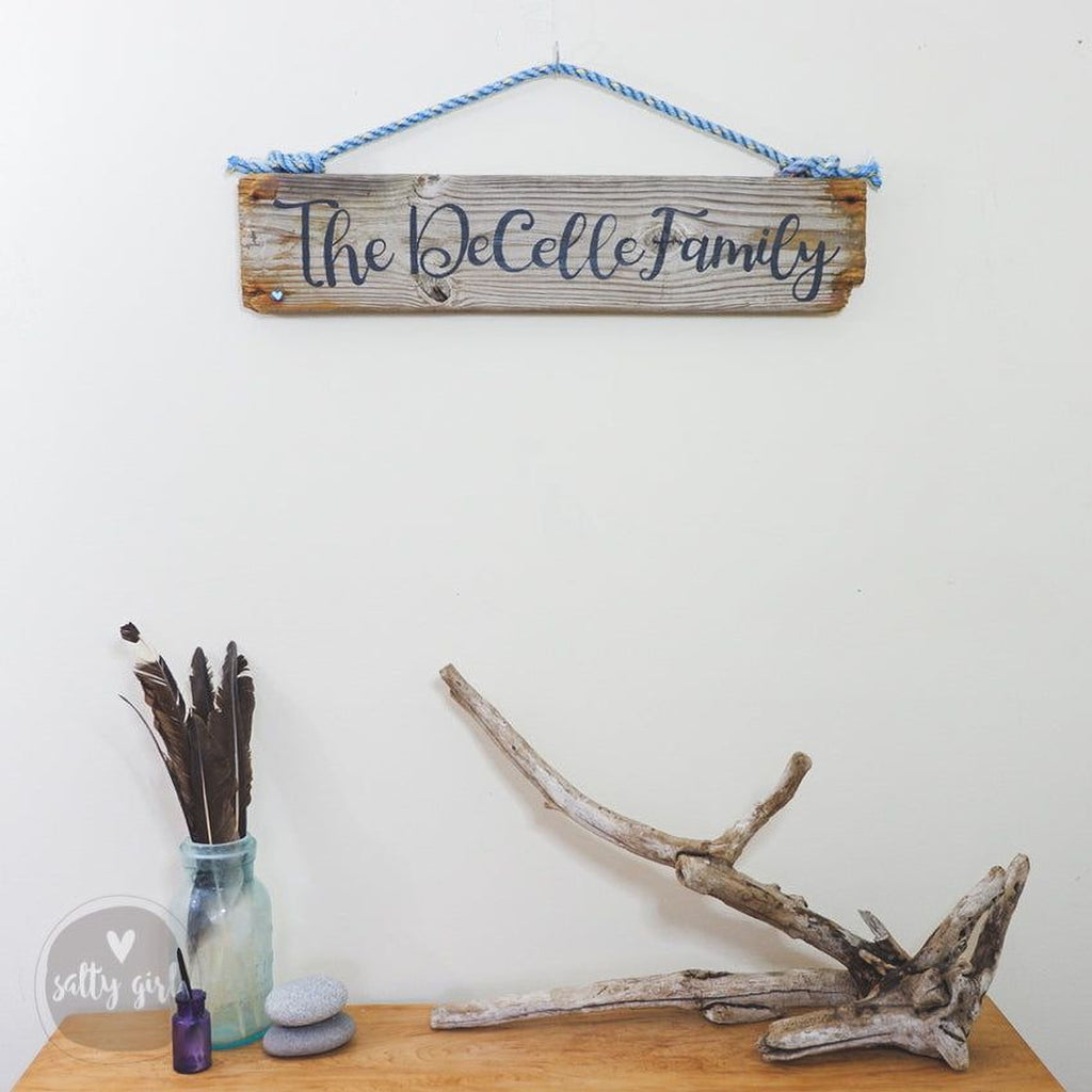 a wooden sign with the name "The Decelle Family" hanging on a white wall, accompanied by a wooden branch, a glass jar with feathers, and a small rock on a wooden surface.