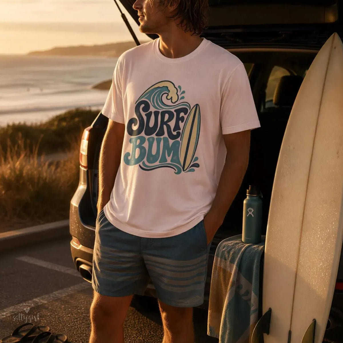 A man stands next to a car, wearing a white t-shirt with a graphic design and holding a surfboard, with a towel and water bottle nearby.