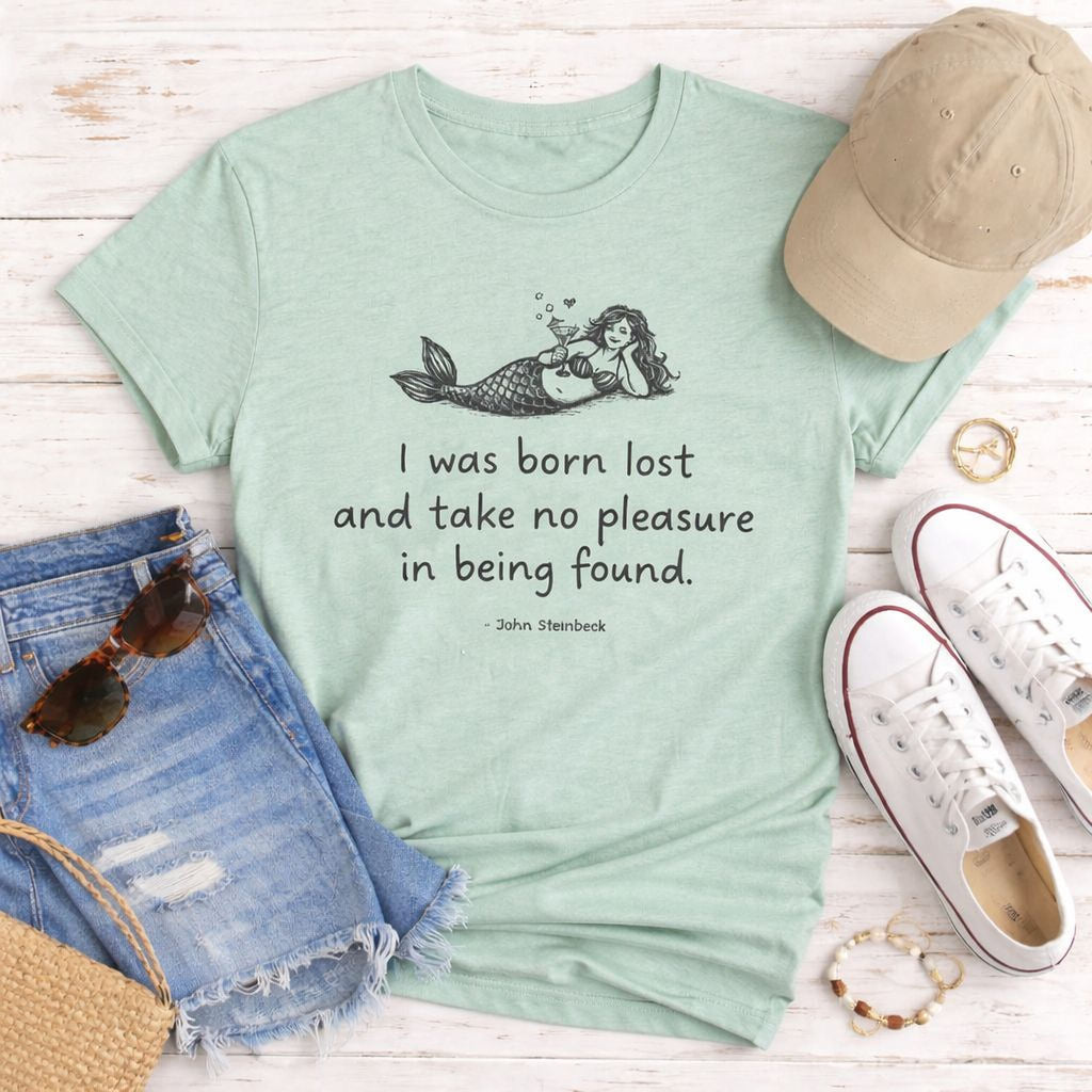 A mint green Maine Salty Girl t-shirt with a mermaid graphic and a quote, a pair of denim shorts, a beige baseball cap, a pair of white sneakers, and some accessories such as sunglasses and bracelets, all laid out on a wooden surface.