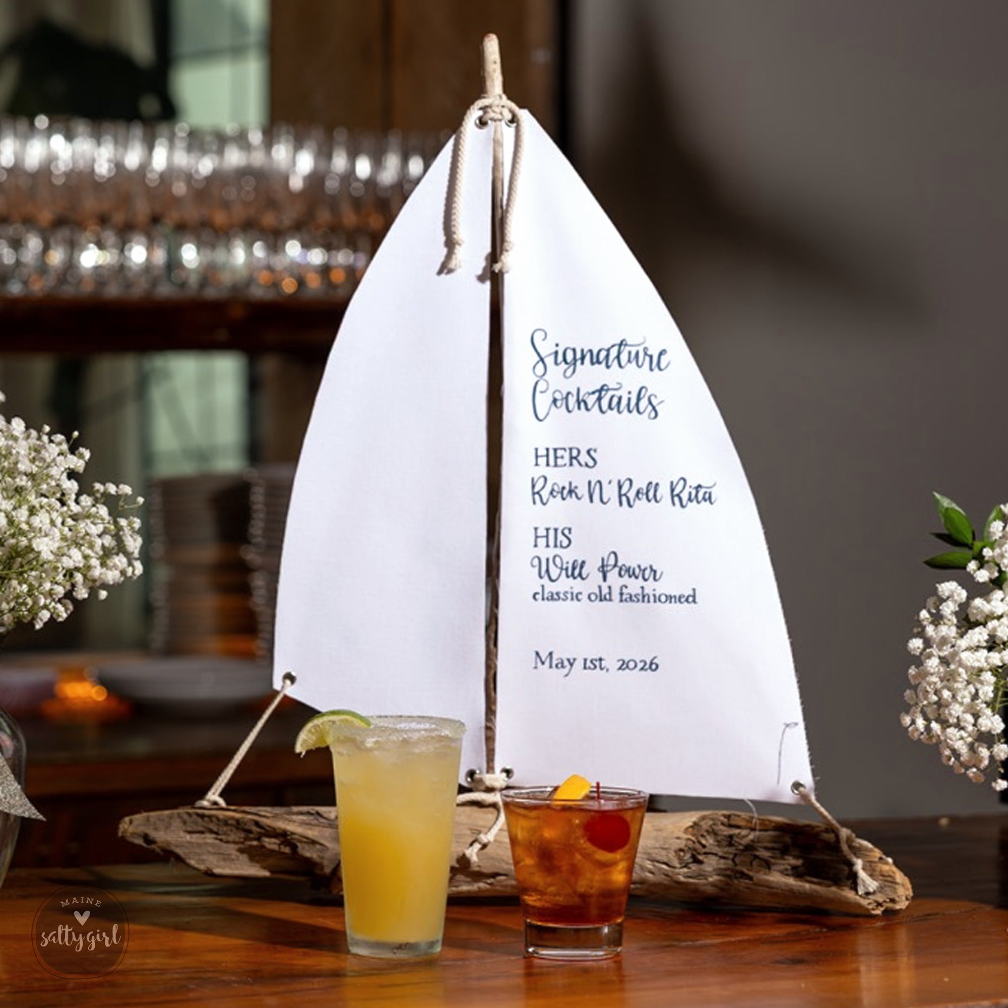 a white sail-shaped cocktail menu with a variety of signature cocktails listed, including "HERS Rock N' Roll Rita", "HIS Will Power", and "Classic Old Fashioned". Two signature cocktails are displayed on a wooden table, one in a tall glass and the other in a short glass. The background includes a blurred view of a bar or restaurant setting with a chandelier and other decor.