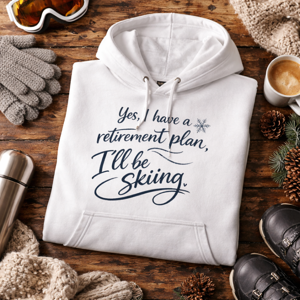 A white hoodie with a humorous retirement plan printed on it, along with ski gear and accessories, such as gloves, a cup, and a pair of shoes, all arranged on a wooden surface.