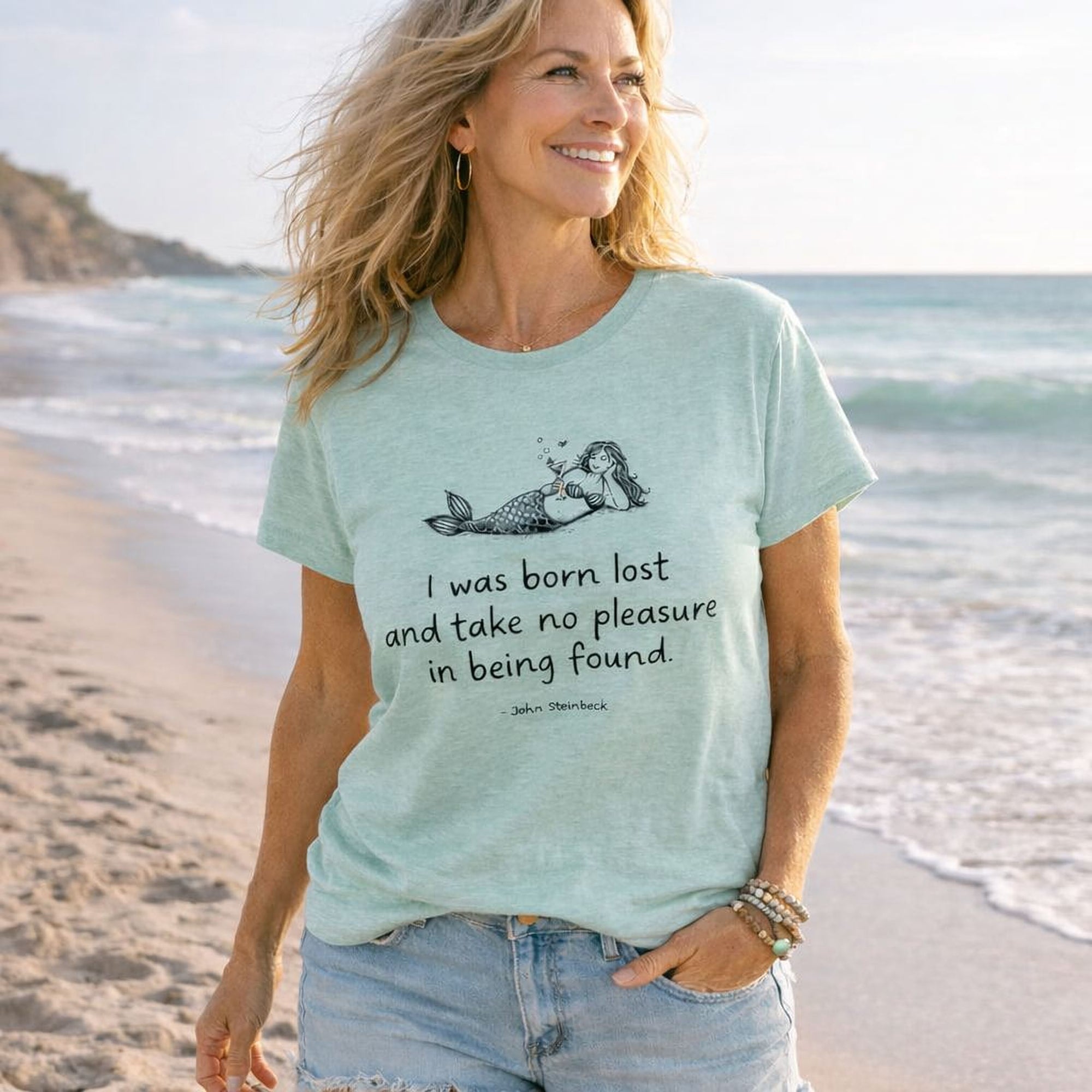A smiling woman wearing a light blue Maine Salty Girl t-shirt with a mermaid graphic and text, standing on a beach with the ocean in the background.