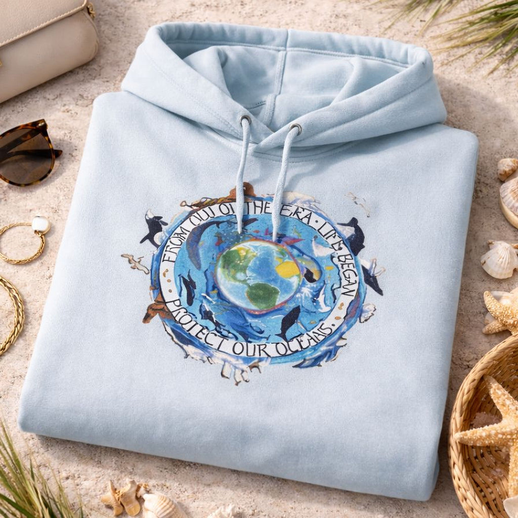 A light blue Maine Salty Girl hoodie with a graphic design of a globe surrounded by marine life, including dolphins and a message about protecting the oceans, is displayed on a beach setting with various accessories and natural elements.