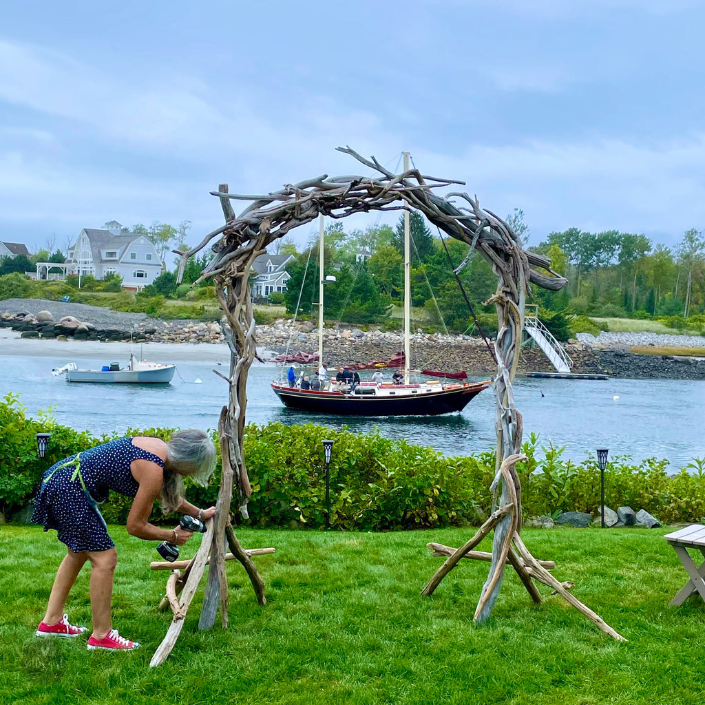 A woman is working on a wooden structure in a grassy area, with a sailboat visible in the background.