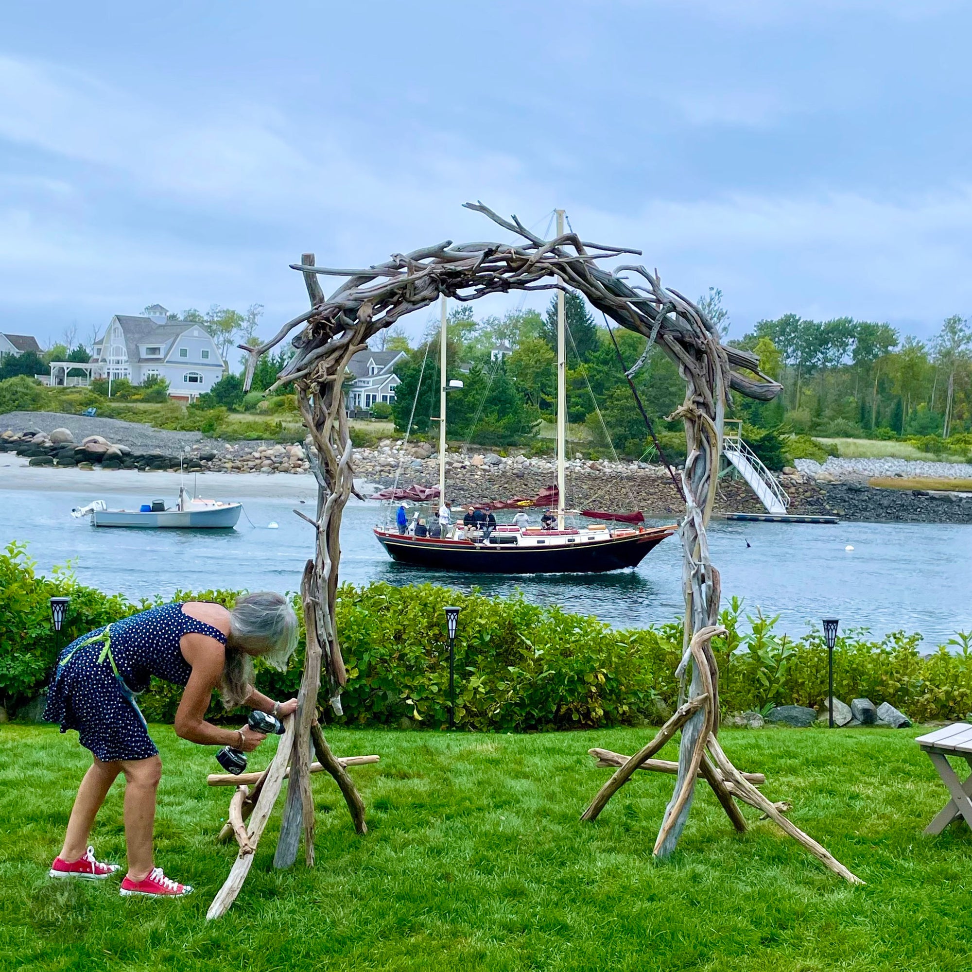 A woman is working on a wooden structure in a grassy area, with a sailboat visible in the background.