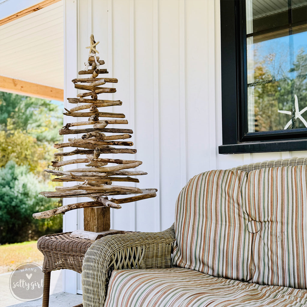 A rustic driftwood Christmas tree stands on a wicker table next to a striped couch, with a white building and trees in the background.