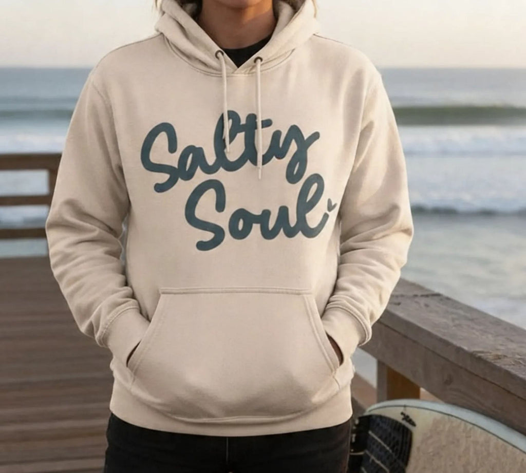 A young woman wearing a beige Maine Salty Girl hoodie with the text "Salt Soul" stands on a wooden pier overlooking the ocean.