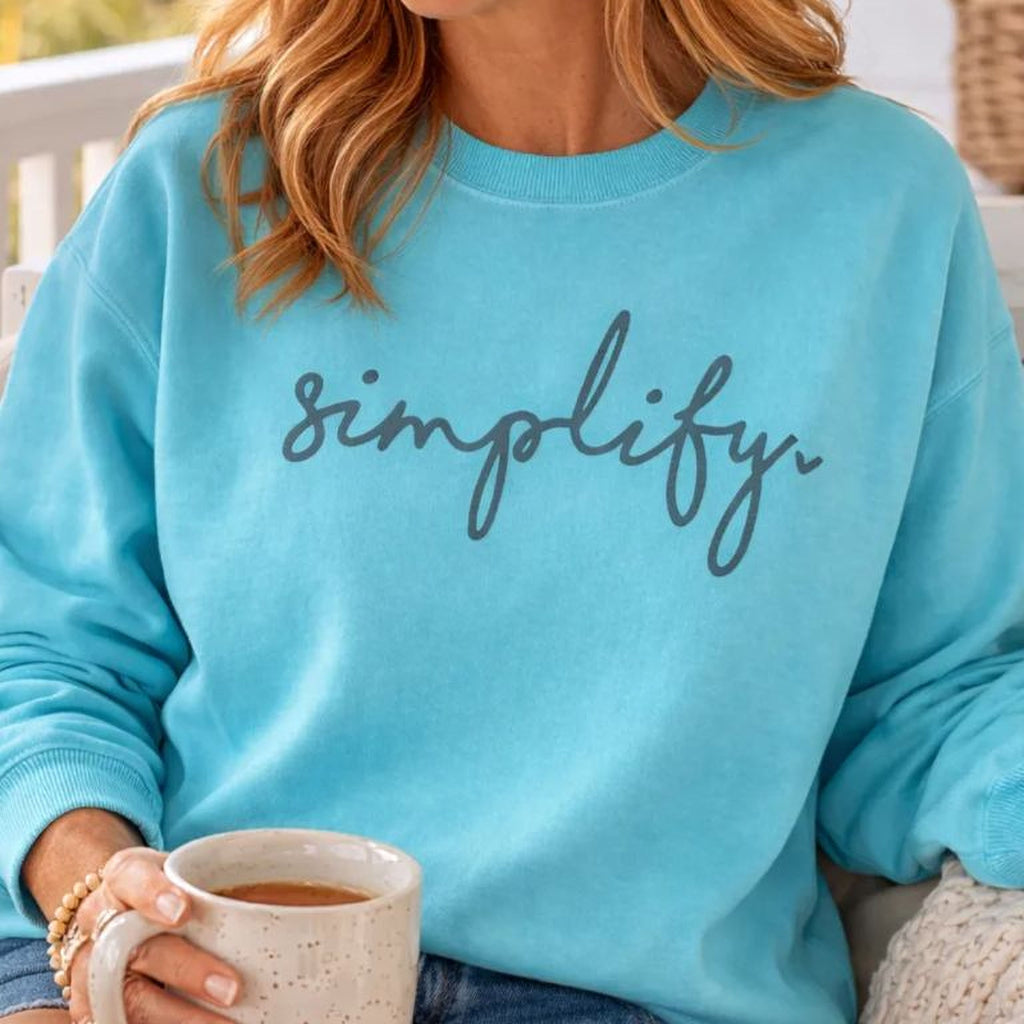 A woman with long, wavy blonde hair is sitting on a porch swing, holding a coffee mug and smiling. She is wearing a blue Maine Salty Girl sweatshirt with the word "simplify" printed on it.