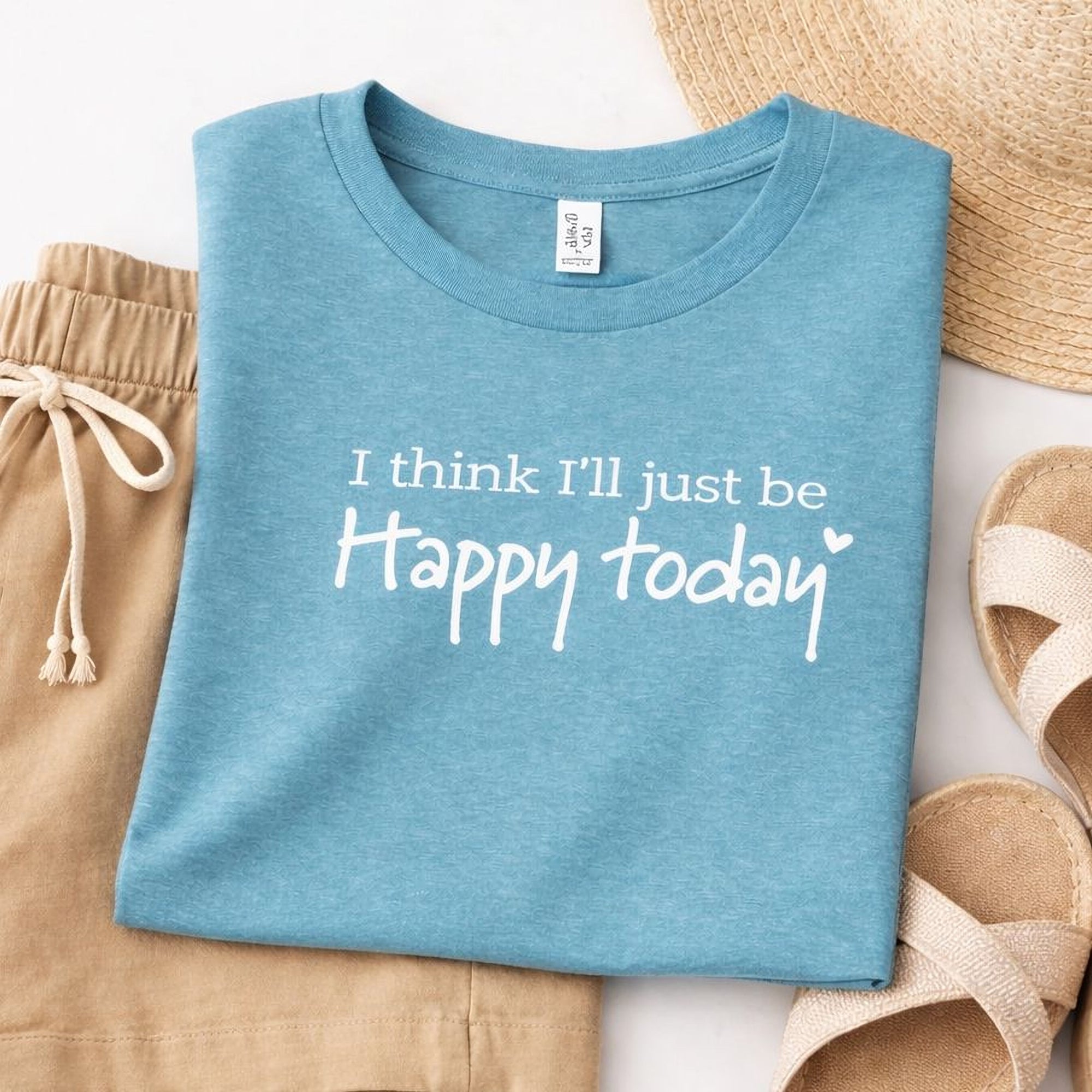 A blue t-shirt Maine Salty Girl with the text "I think I'll just be happy today" printed on it, along with a straw hat and sandals, all laid out on a white background.