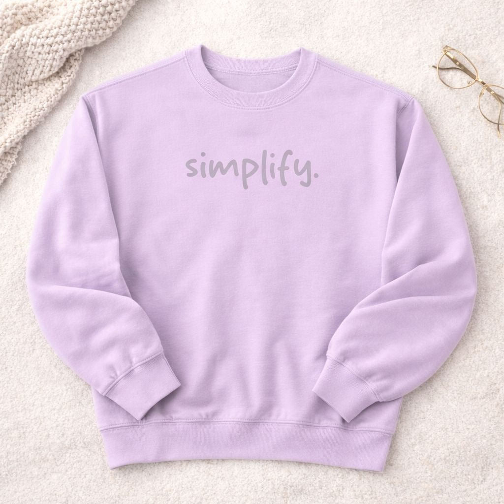 A lavender-colored sweatshirt with the word "simplify" printed on it, placed on a white surface next to a pair of glasses.