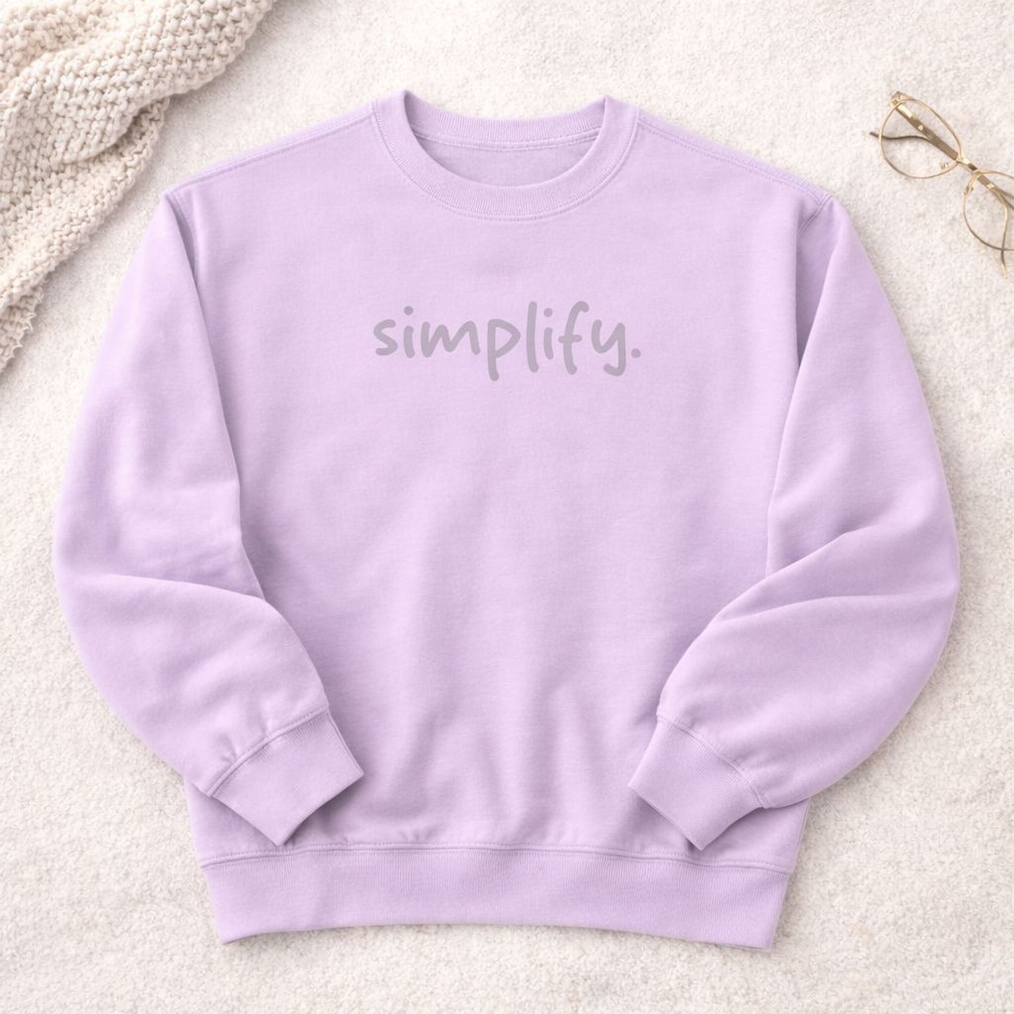 A lavender-colored sweatshirt with the word "simplify" printed on it, placed on a white surface next to a pair of glasses.