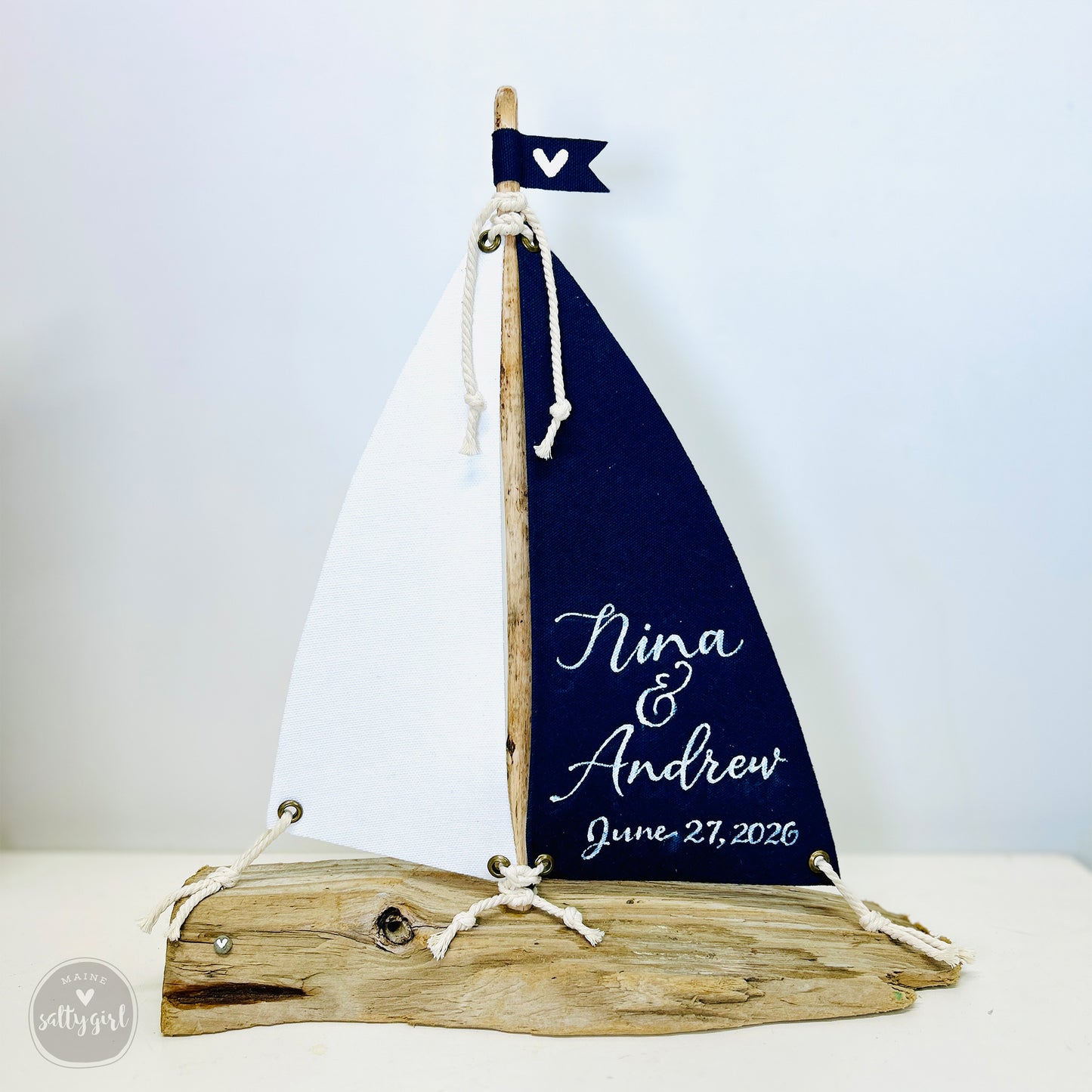 A wooden sailboat-shaped wedding decoration with a white sail and a blue sail, featuring the names 'Nina & Andrew' and the date 'June 27, 2026'.