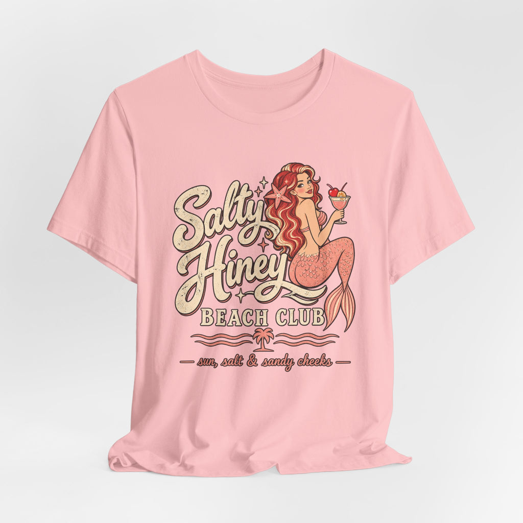 A pink t-shirt with a graphic design featuring a mermaid character and text that says "Salt & Honey Beach Club".