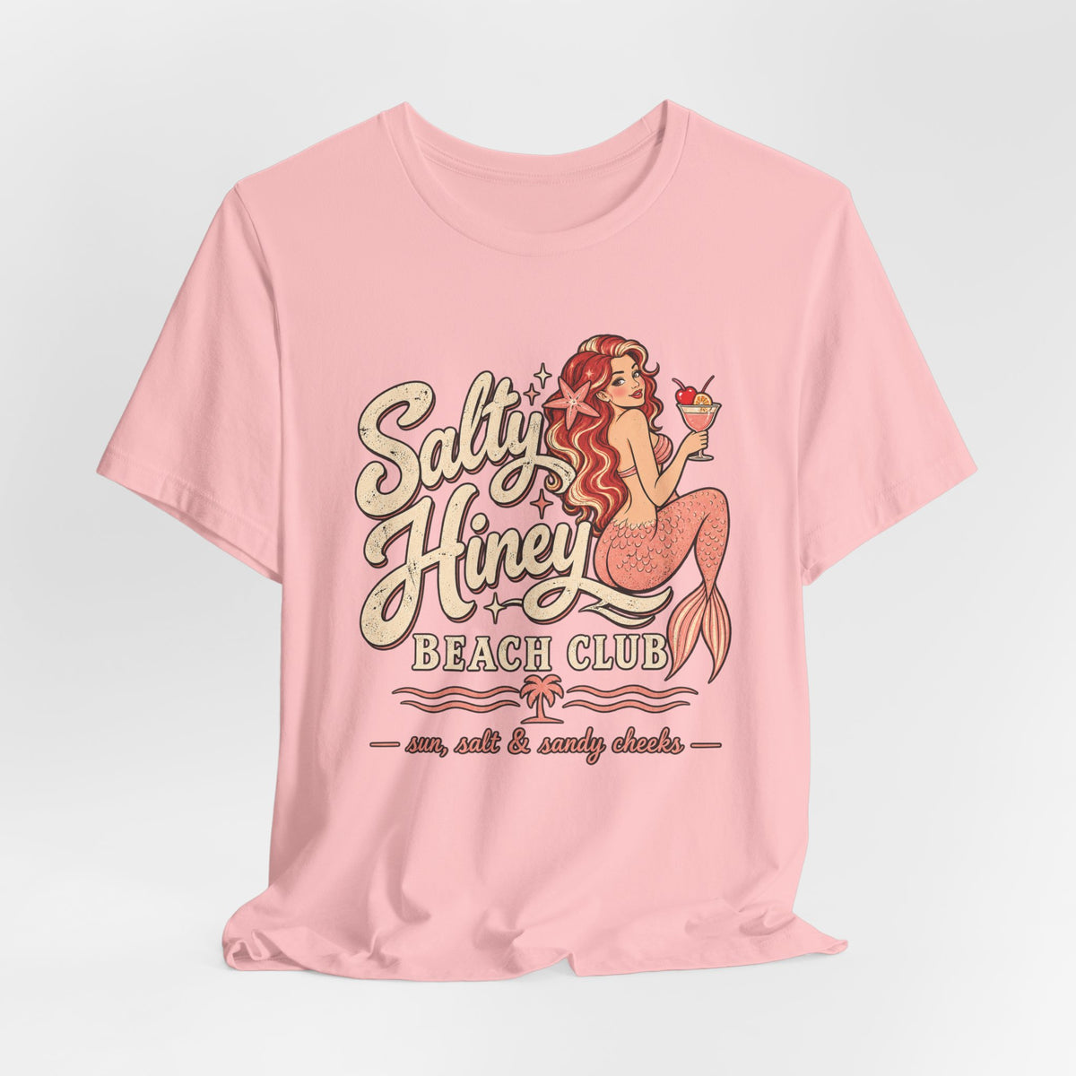 A pink t-shirt with a graphic design featuring a mermaid character and text that says "Salt & Honey Beach Club".