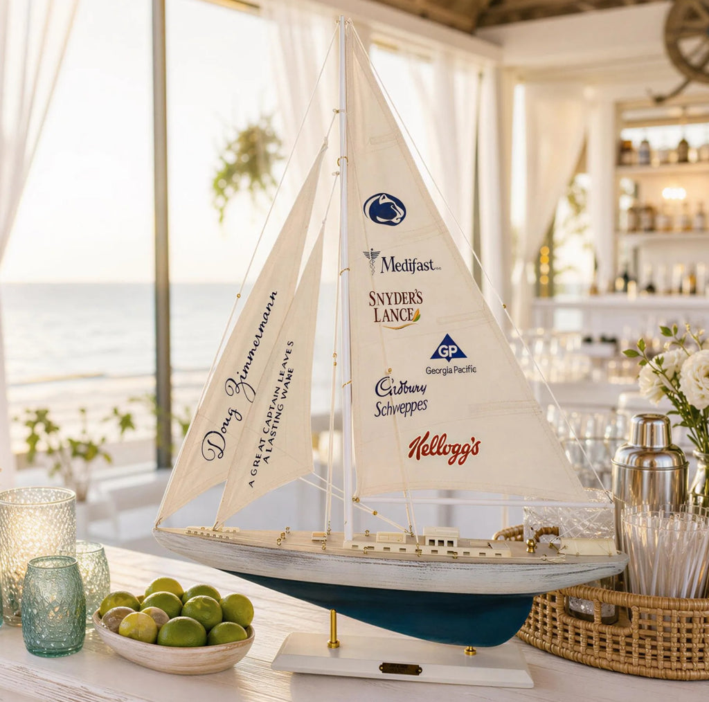 A model sailboat with sail and hull decorations sits on a table, surrounded by various items such as a bowl of fruit, a vase, and other decorative objects.