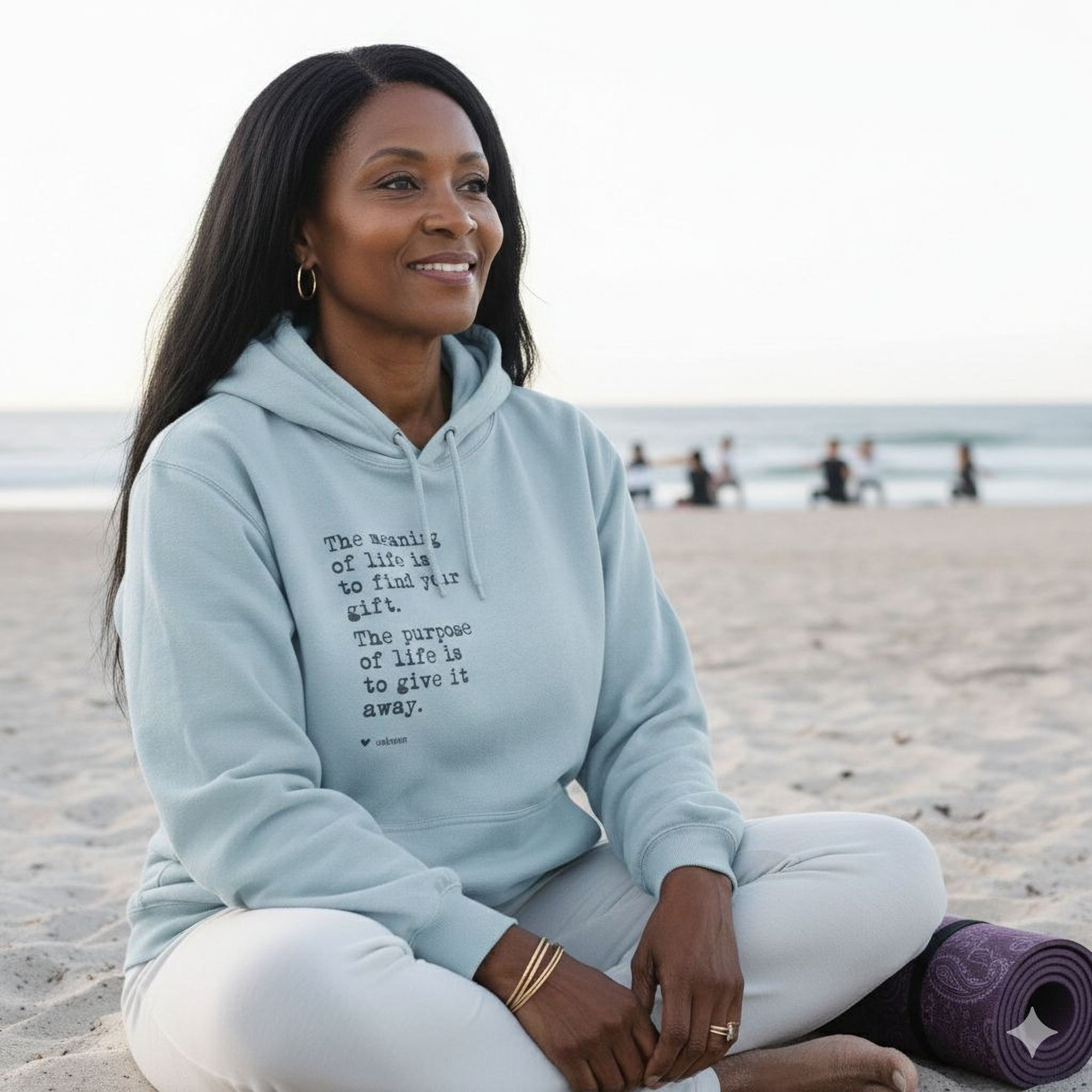 A woman in a light blue Maine Salty Girl hoodie sits on a sandy beach, smiling and looking towards the ocean.