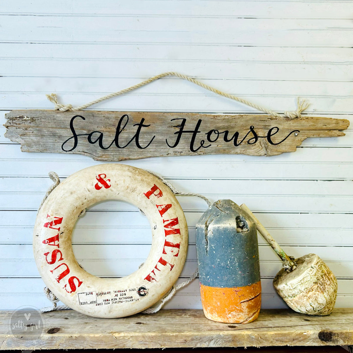 a rustic wooden sign that reads "Salt House" hanging on a white wall, along with a white life preserver and a blue and orange buoy.