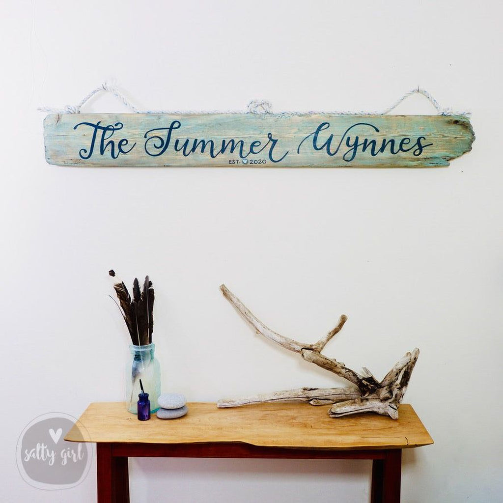 a wooden sign with the name "The Summer Wynn" hanging on a wall above a wooden table with various items on it, including a vase with feathers and a bottle.