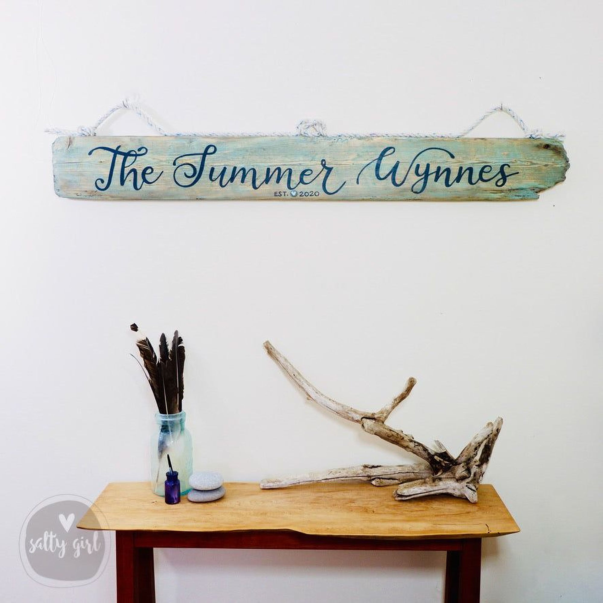 a wooden sign with the name "The Summer Wynn" hanging on a wall above a wooden table with various items on it, including a vase with feathers and a bottle.