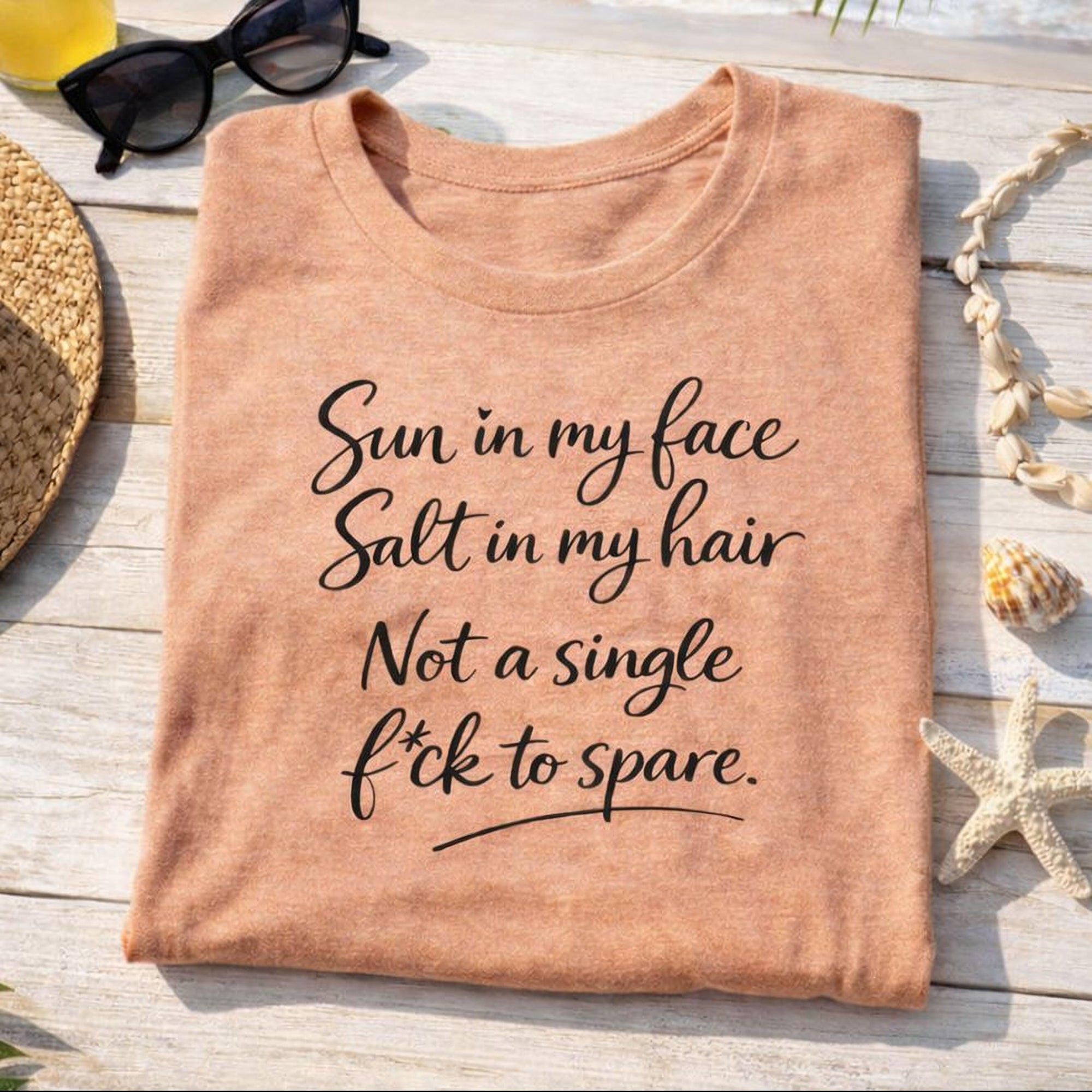 A Maine Salty Girl t-shirt with a humorous text printed on it, placed on a wooden surface with a straw hat, sunglasses, and seashells nearby.
