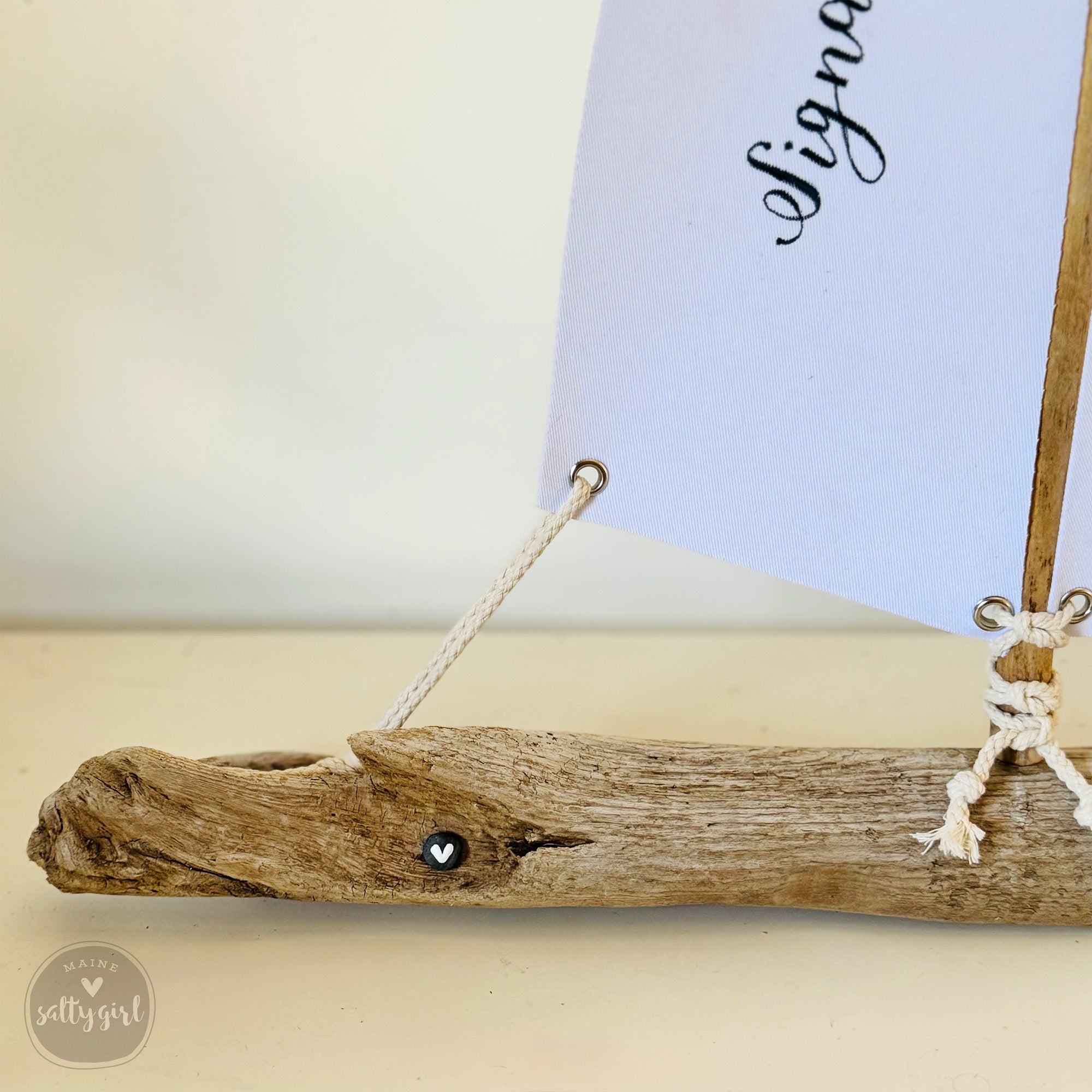 A small driftwood boat with a white sail attached to it, sitting on a white surface.