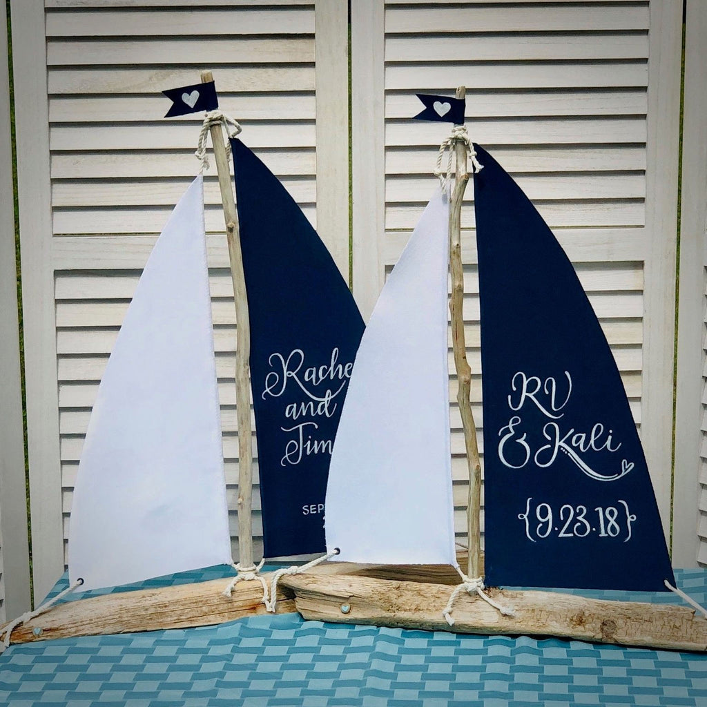 two wooden sailboat-shaped wedding decorations, each with a white sail and a heart-shaped flag. The sailboats are placed on a blue and white checkered tablecloth, and the names "Rachael and Tim" are written on the sails, indicating the names of the couple.