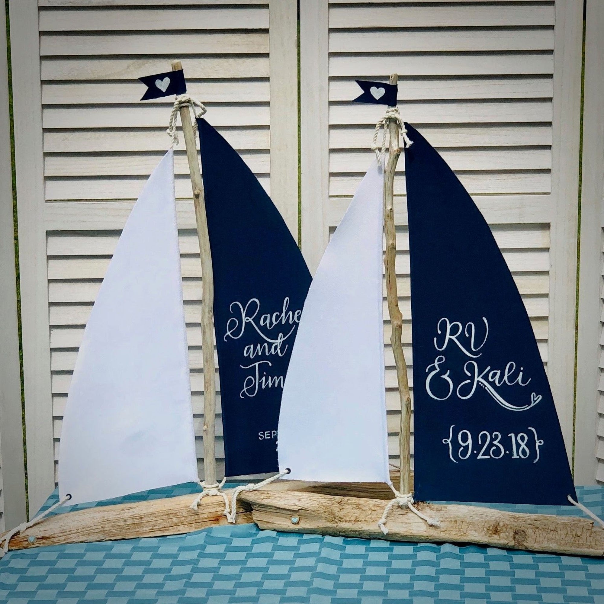 two wooden sailboat-shaped wedding decorations, each with a white sail and a heart-shaped flag. The sailboats are placed on a blue and white checkered tablecloth, and the names "Rachael and Tim" are written on the sails, indicating the names of the couple.