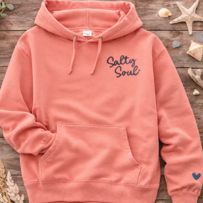 A pink hoodie with the text "Salty Soul" printed on it, along with a heart symbol on the left sleeve, is displayed on a wooden surface alongside various seashells, starfish, and a plant.