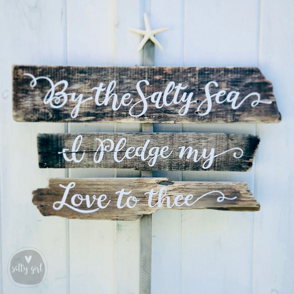 A wooden sign with the text "By the Salty Sea" and "Ipledge my Love to thee" written on it, mounted on a white wooden wall.