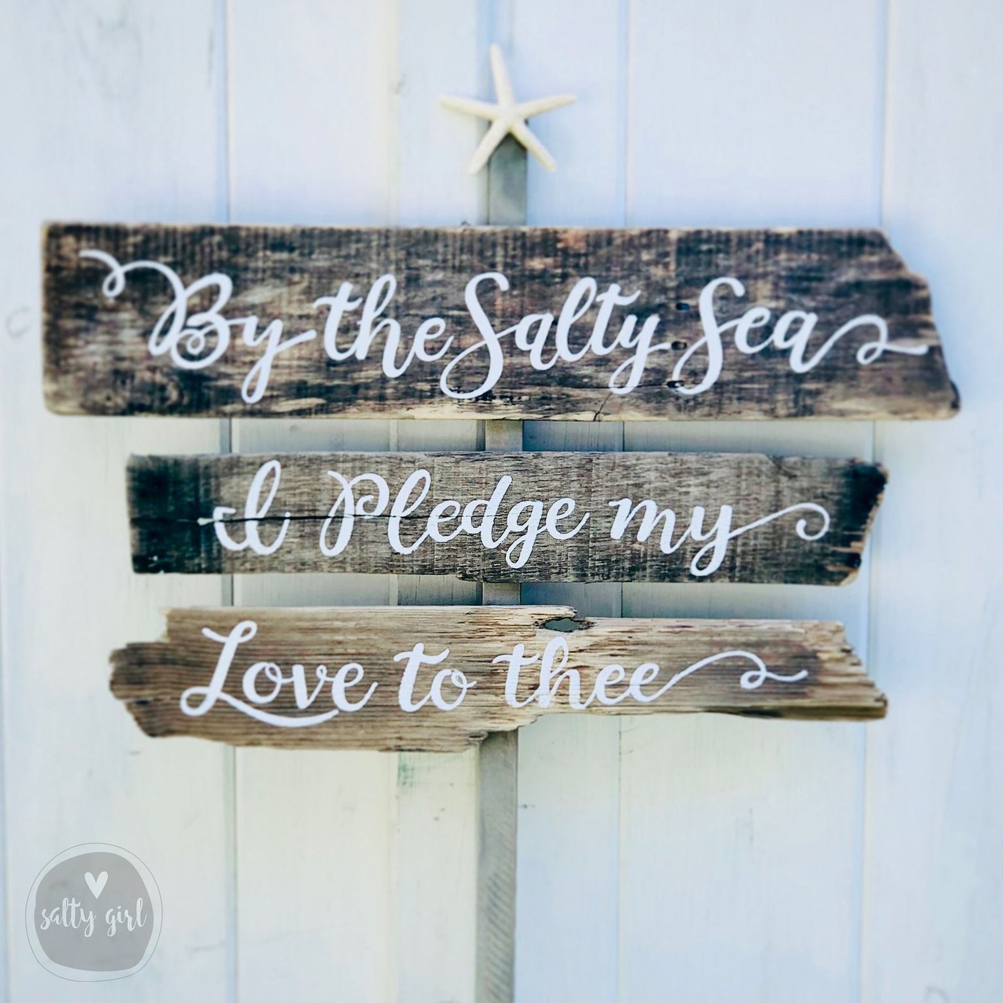 A wooden sign with the text "By the Salty Sea" and "Ipledge my Love to thee" written on it, mounted on a white wooden wall.