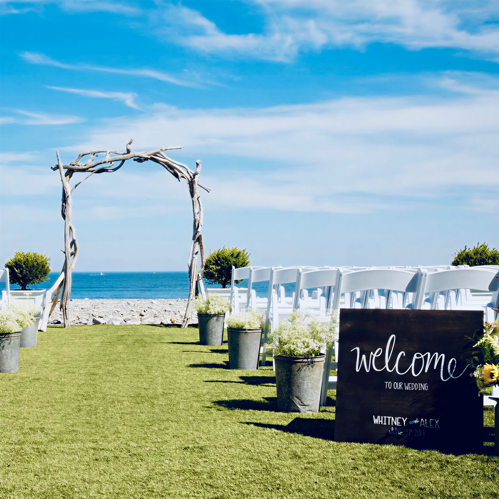 a beautiful outdoor wedding ceremony setup with a wooden arch, white chairs, and potted plants arranged on a grassy lawn overlooking the ocean.