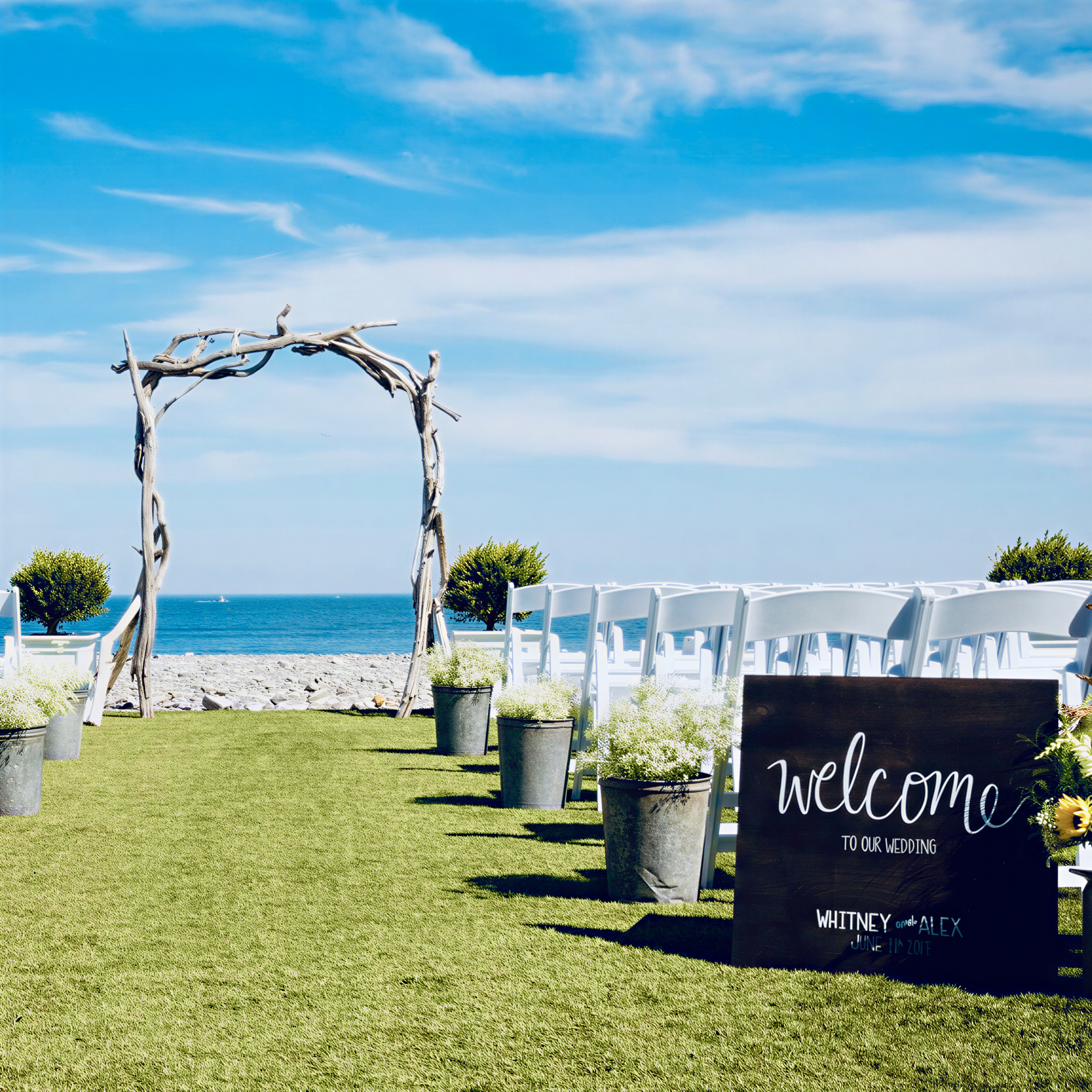 a beautiful outdoor wedding ceremony setup with a wooden arch, white chairs, and potted plants arranged on a grassy lawn overlooking the ocean.