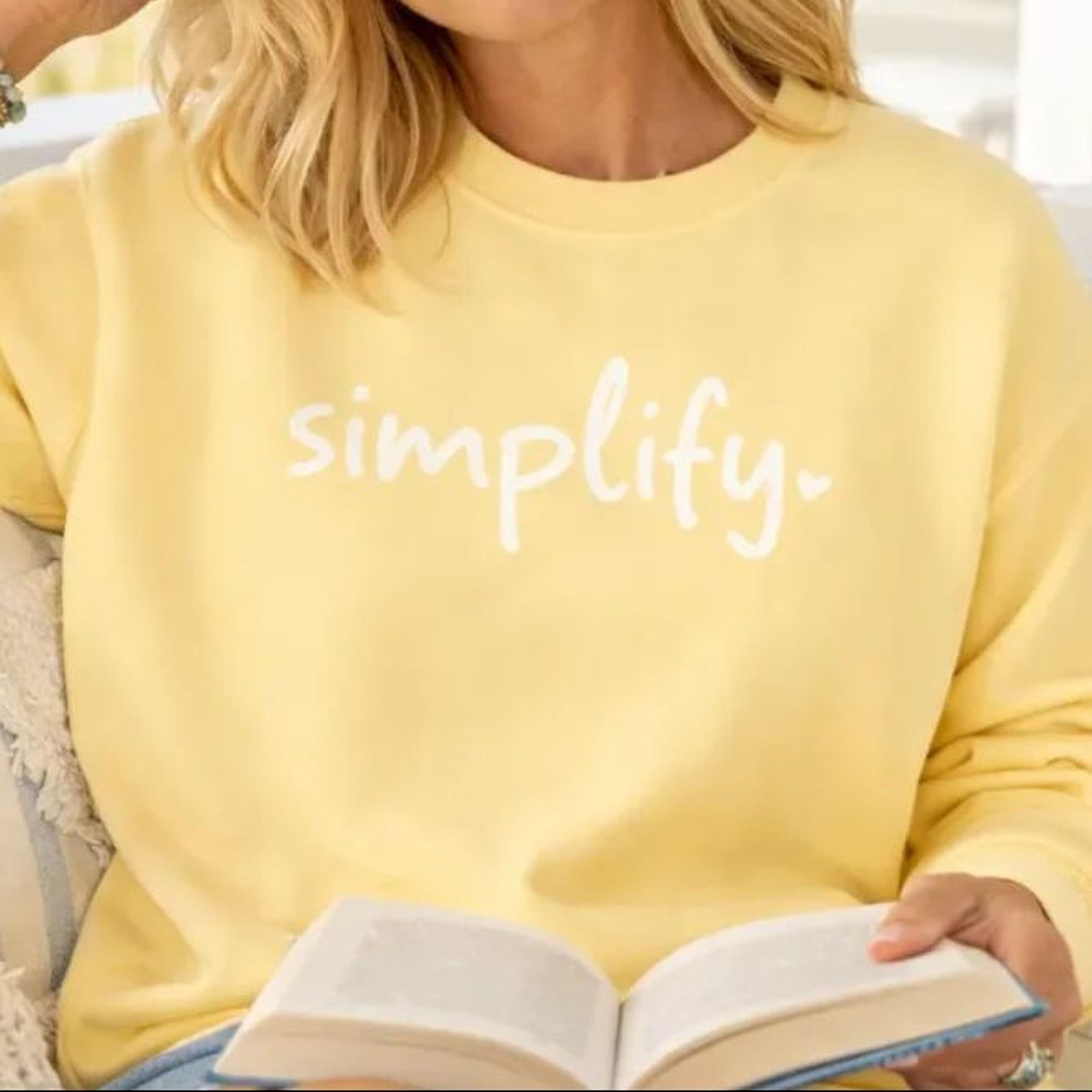 A woman with blonde hair is sitting on a bench, reading a book and wearing a yellow Maine Salty Girl sweatshirt with the word "simplify" printed on it.