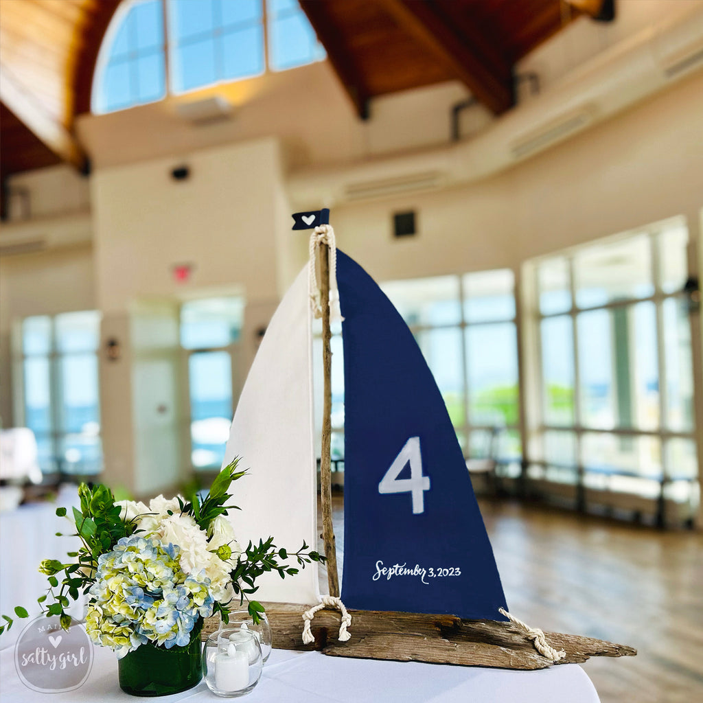 A decorative table with a blue sailboat-shaped sign displaying the number 4, a vase of white and blue flowers, and candles, set against a blurred background of a large room with windows.