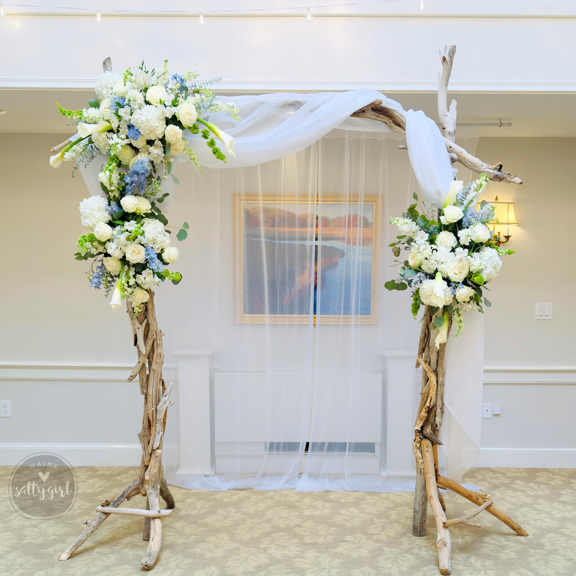 A beautifully decorated wedding arch made of driftwood and adorned with white and blue flowers, standing in front of a painting.