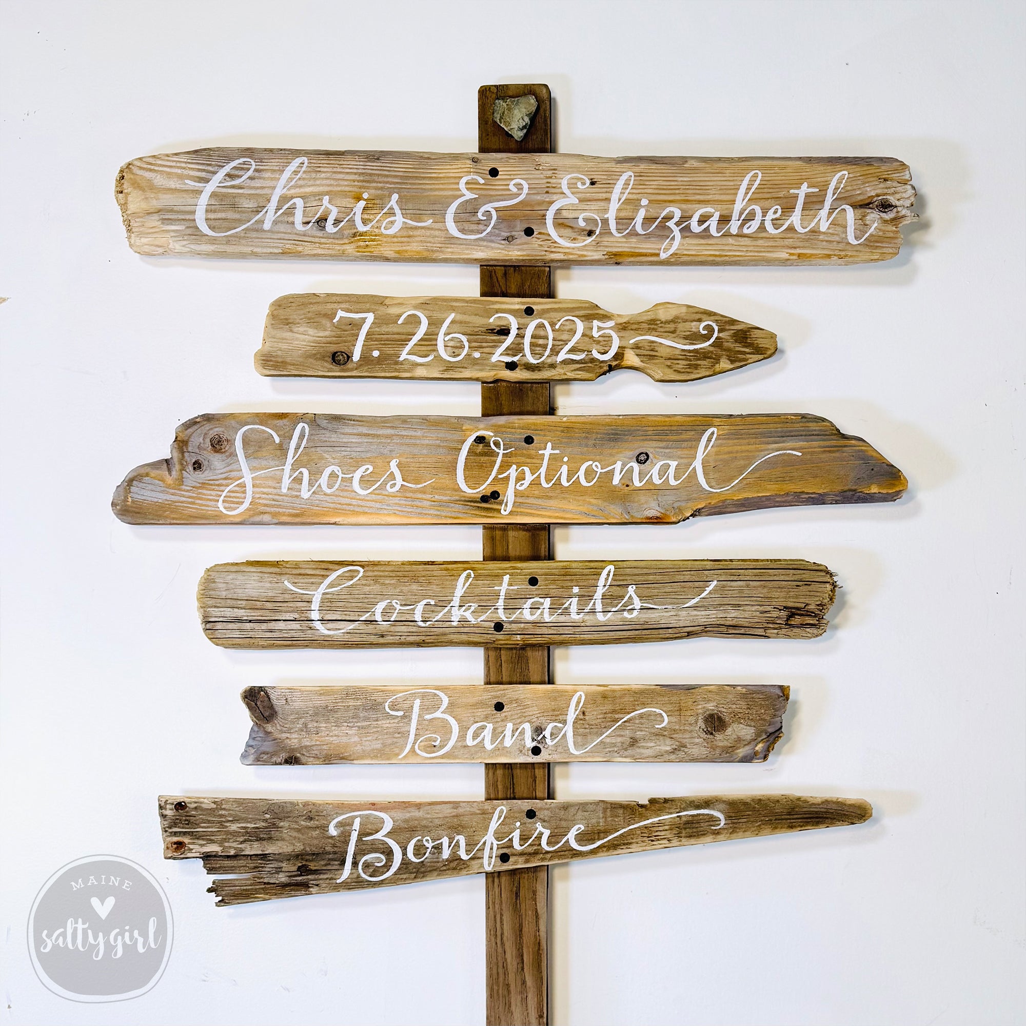 A wooden sign with the names "Chris & Elizabeth" and the dates "7.26.2025" displayed on it, along with additional text indicating "Shoes Optional", "Cocktails", "Band", and "Bonfire".