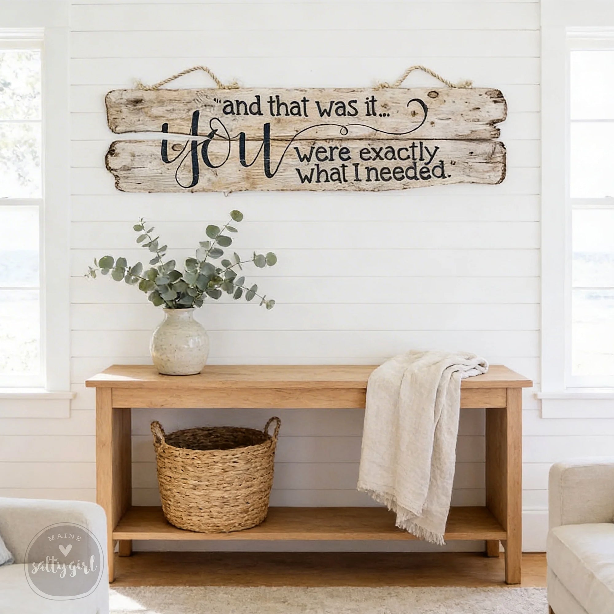 a rustic wooden wall sign with a quote, a woven basket, and a vase of greenery, all set against a white wall with a window.