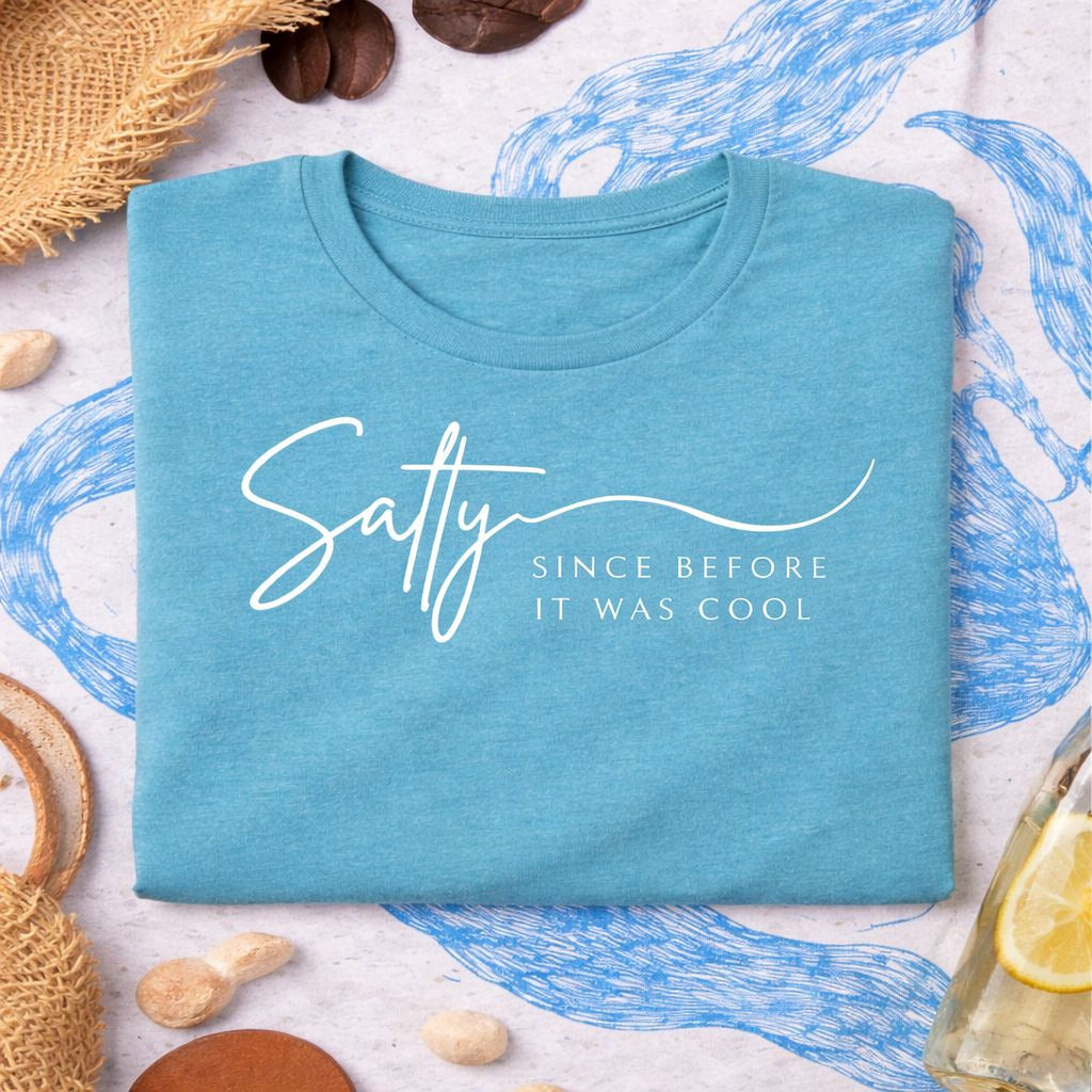 a blue t-shirt with the text "Salty Since before it was cool" printed on it, along with some other items such as a straw hat, a lemon slice, and some coffee beans.