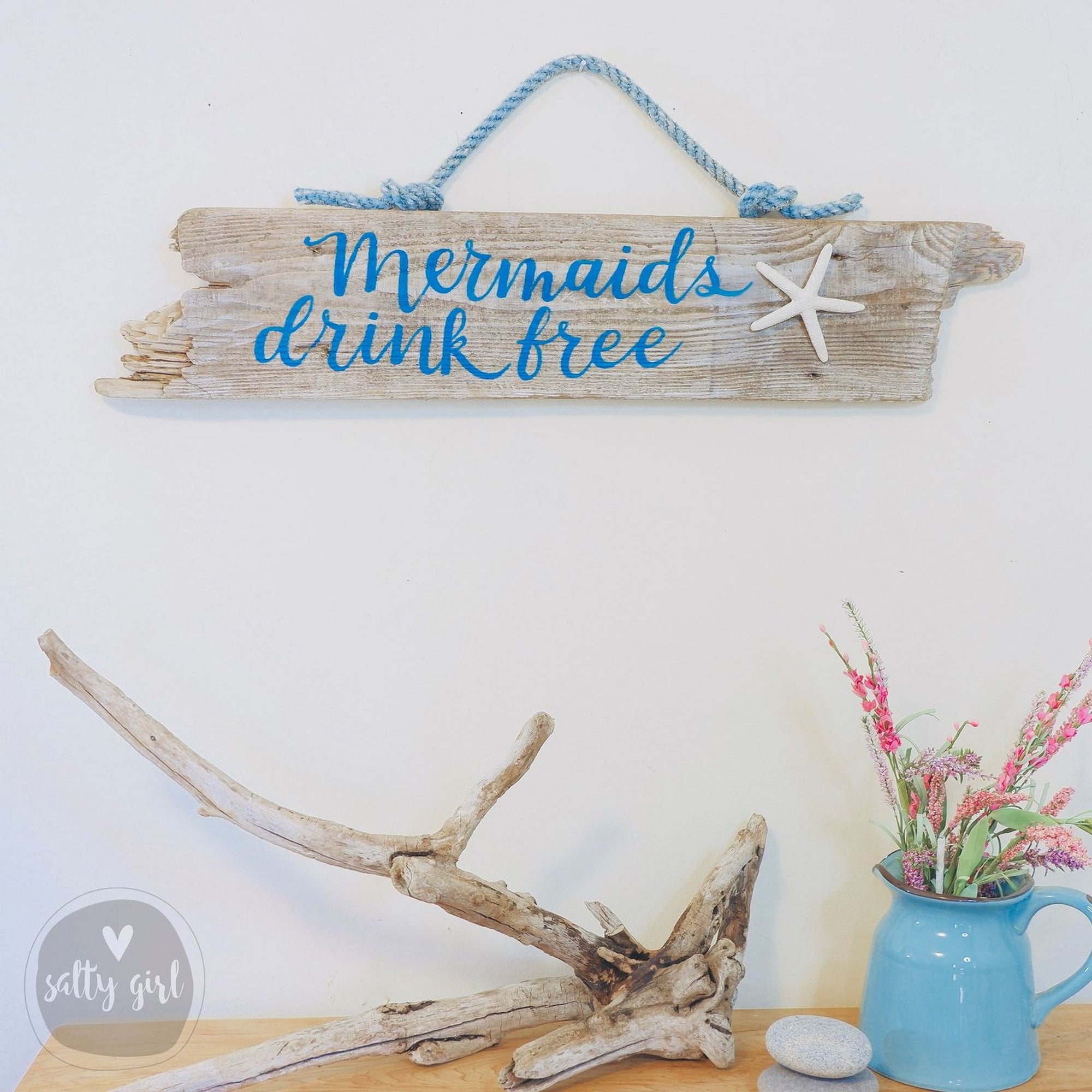 a wooden sign with the text "Mermaids drink free" written on it, hanging on a wall. Below the sign, there is a blue vase with pink flowers, and a wooden branch with a starfish on it. The overall scene has a coastal, nautical theme.