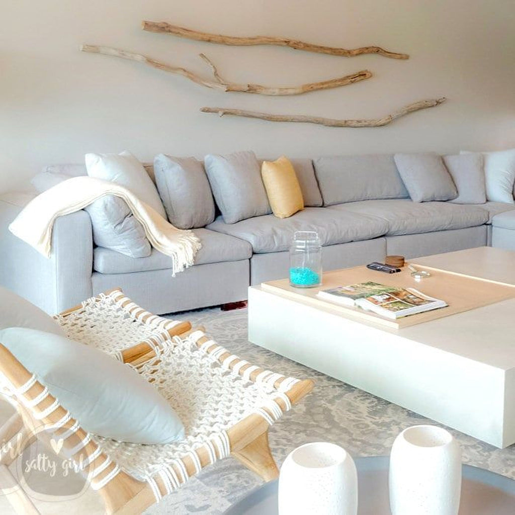 a cozy living room with a gray sectional sofa, a woven chair, and a coffee table with a magazine and a glass jar. The room has a neutral color palette with pops of color from the throw pillows and the woven chair. The wall features a unique arrangement of driftwood branches, adding a rustic touch to the space.