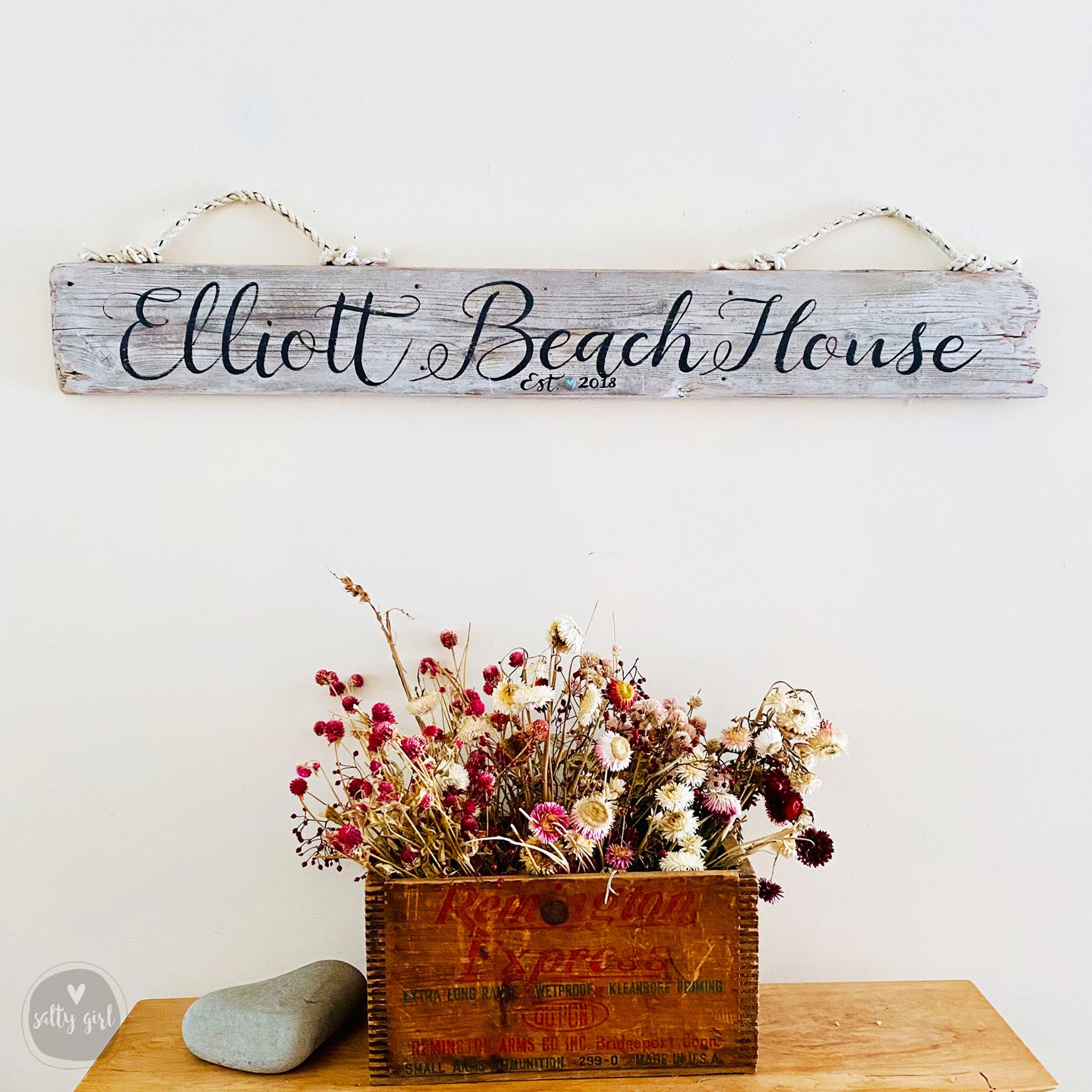a wooden crate filled with dried flowers, placed on a wooden surface. Above the crate, there is a wooden sign with the name "Elliot Beach House" written on it.