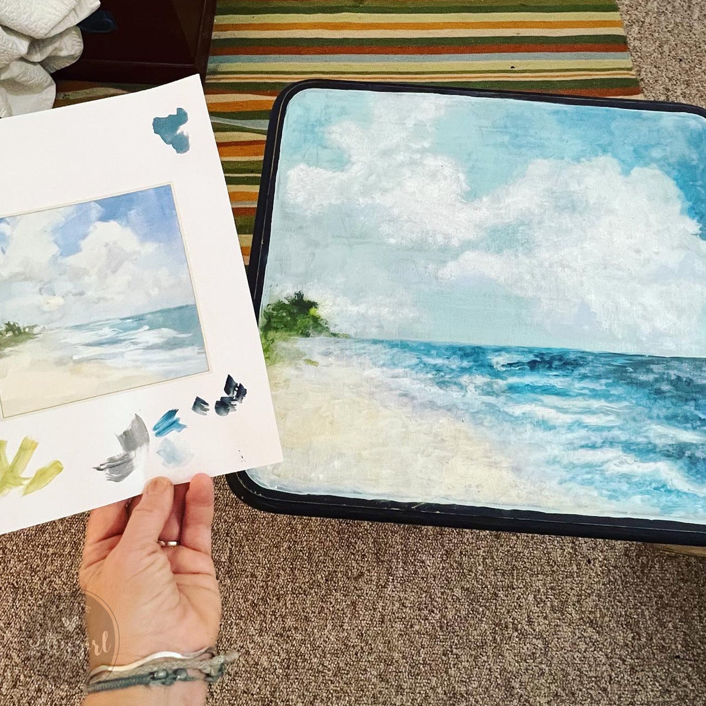 A hand holding a piece of paper with a painting of a beach scene, and another painting of a beach scene on a canvas.