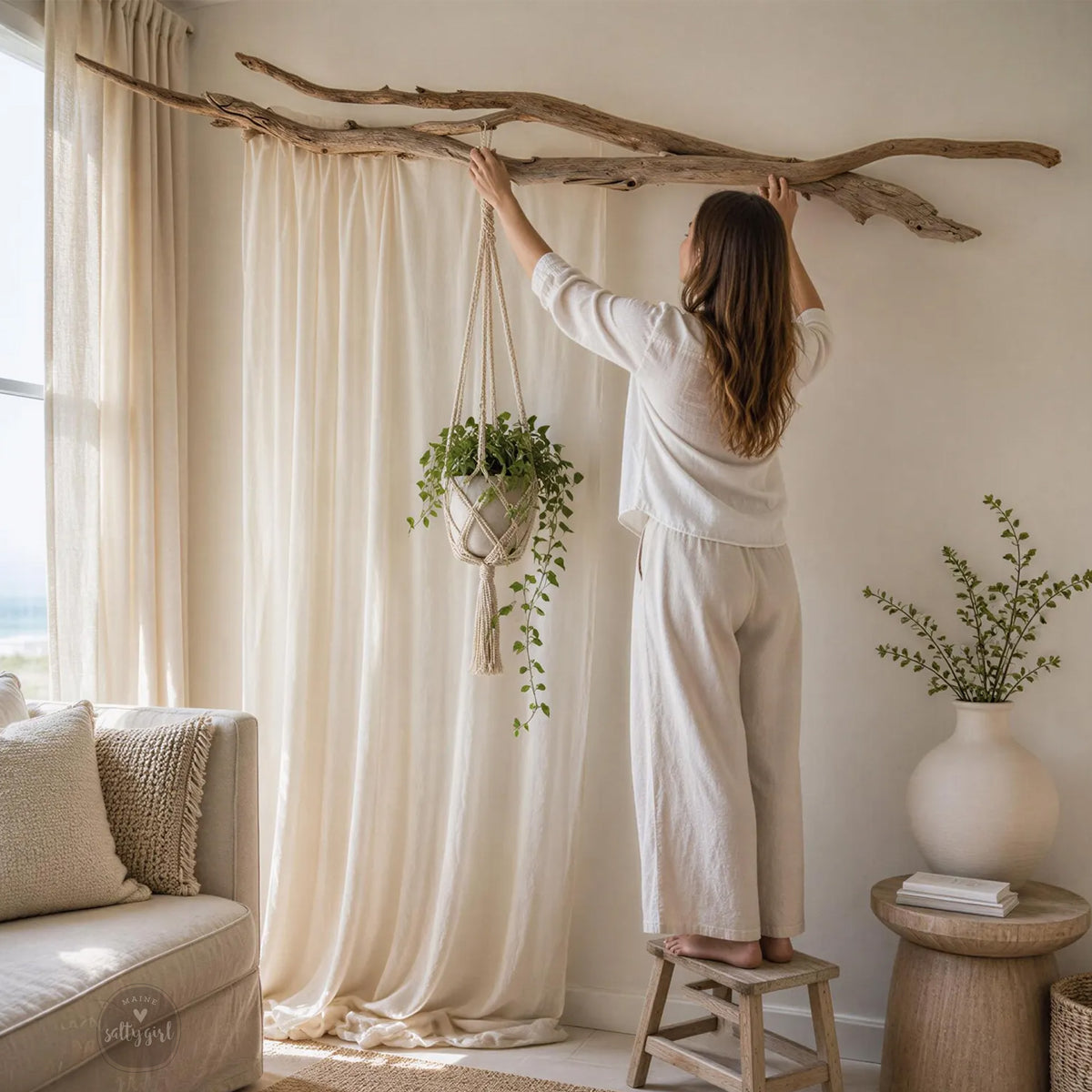 A woman in a white outfit is arranging plants on a wooden stand in a living room with a large branch hanging from the ceiling.