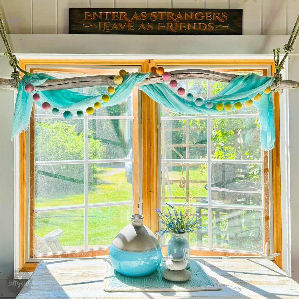 a window with a wooden frame, a blue vase with flowers, and a sign that reads "Enter as strangers, leave as friends". The window is set in a room with a wooden floor and a view of a garden outside.