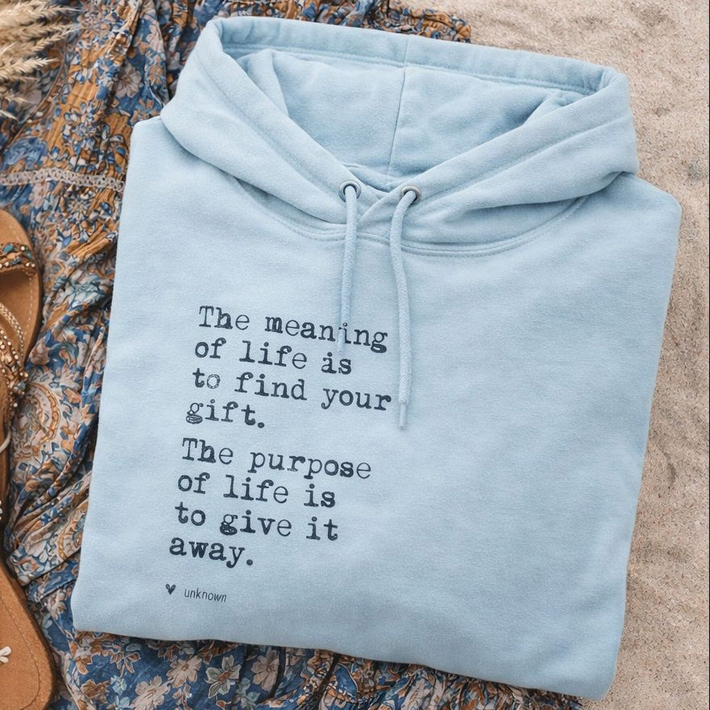 A light blue Maine Salty Girl hoodie with a quote printed on it, laid out on a sandy surface next to a pair of sandals.