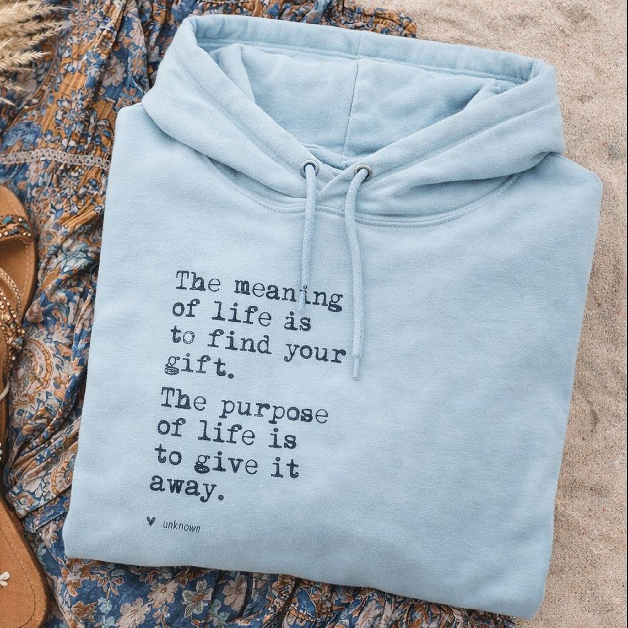 A light blue Maine Salty Girl hoodie with a quote printed on it, laid out on a sandy surface next to a pair of sandals.
