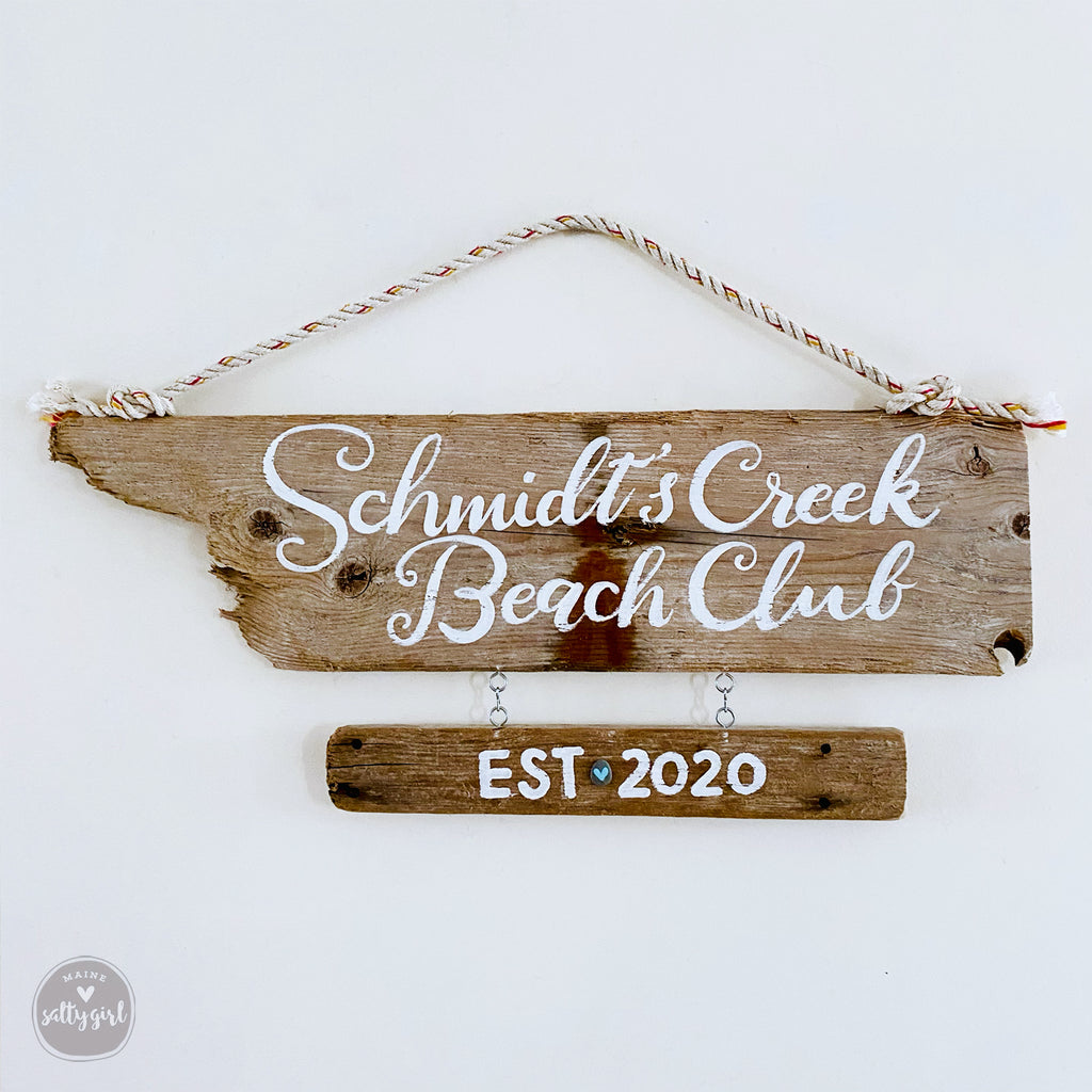 A wooden sign with the text "Schmidt's Creek Beach Club EST 2020" hanging on a wall.