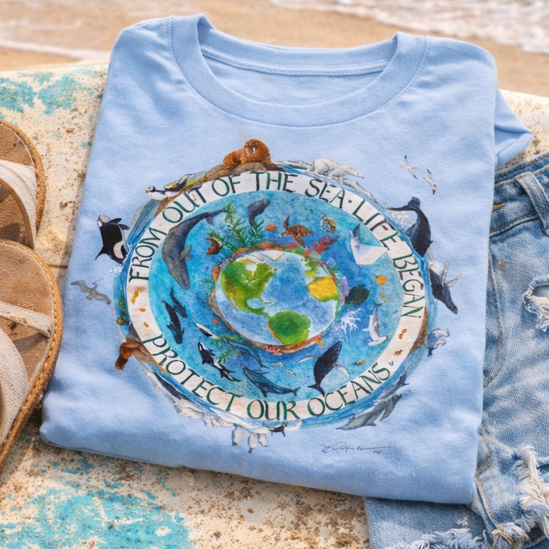 A blue t-shirt with a graphic design of a globe and various marine life, along with a pair of denim shorts and sandals, are placed on a beach towel.
