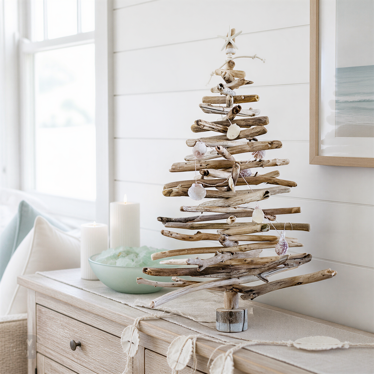 A rustic driftwood Christmas tree stands on a wooden dresser, adorned with seashells and candles, creating a cozy and coastal holiday atmosphere.