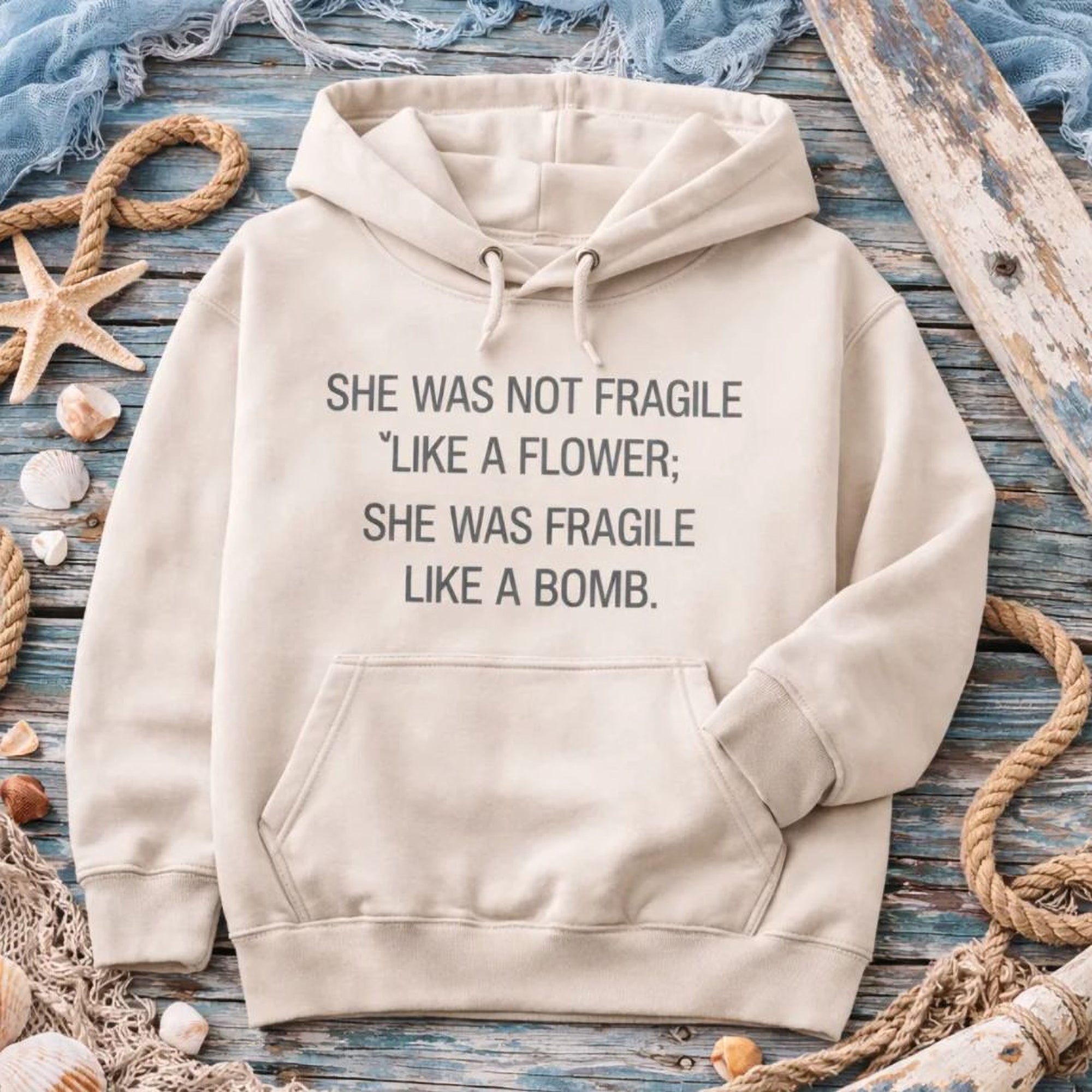 A beige Maine Salty Girl hoodie with a humorous text printed on it, laid out on a wooden surface surrounded by seashells, rope, and starfish.